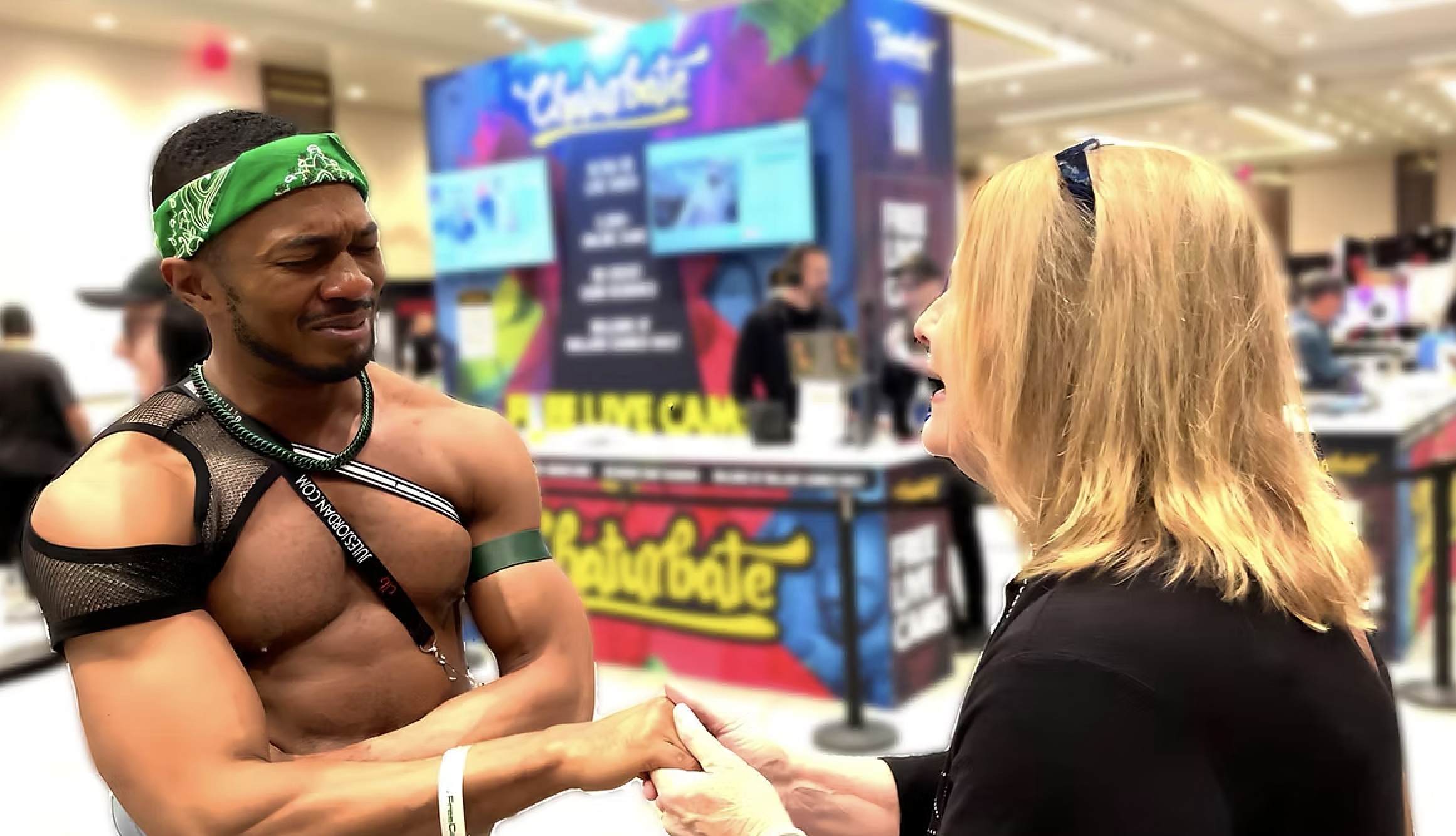 A shirtless man with a green bandana and mesh arm sleeve holding hands with a woman with blond hair, black sunglasses on her head, in conversation at a convention with a colorful background that reads 'Exportate' and a display booth behind them.