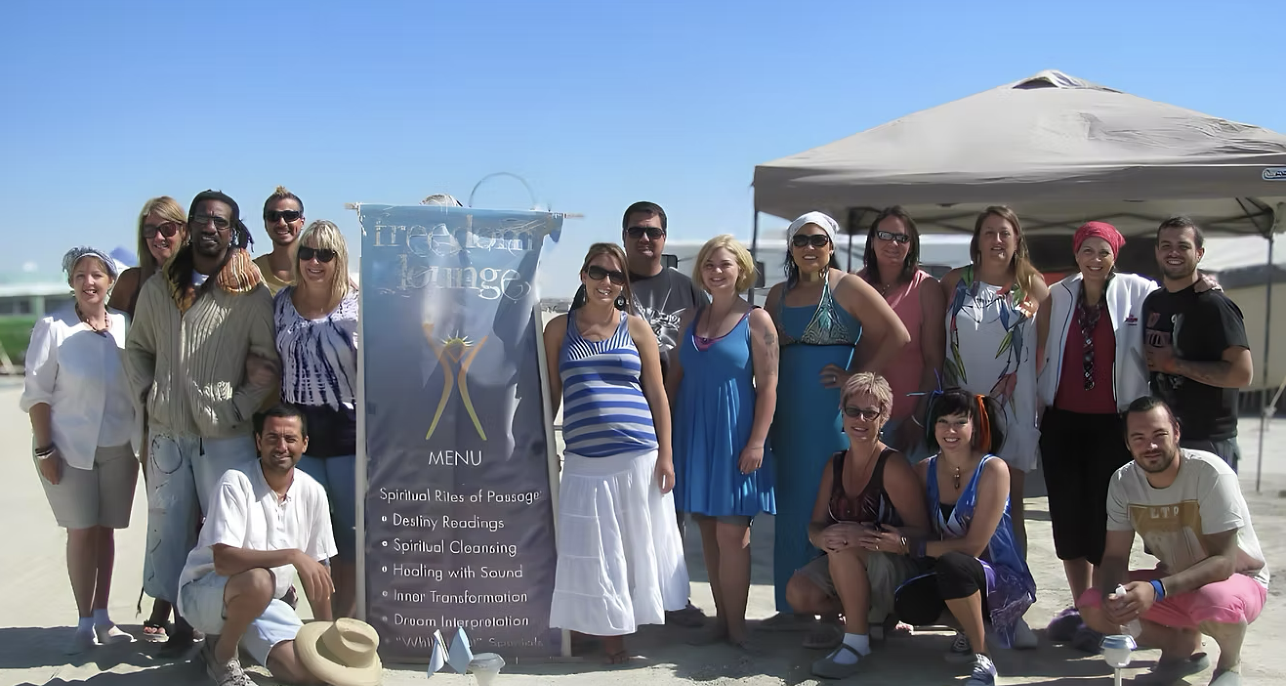 Group of people on a sandy beach, posing for a photo under a canopy with a sign listing spiritual services like readings, cleansing, and sound healing. The sky is clear and blue, with a boat in the background.