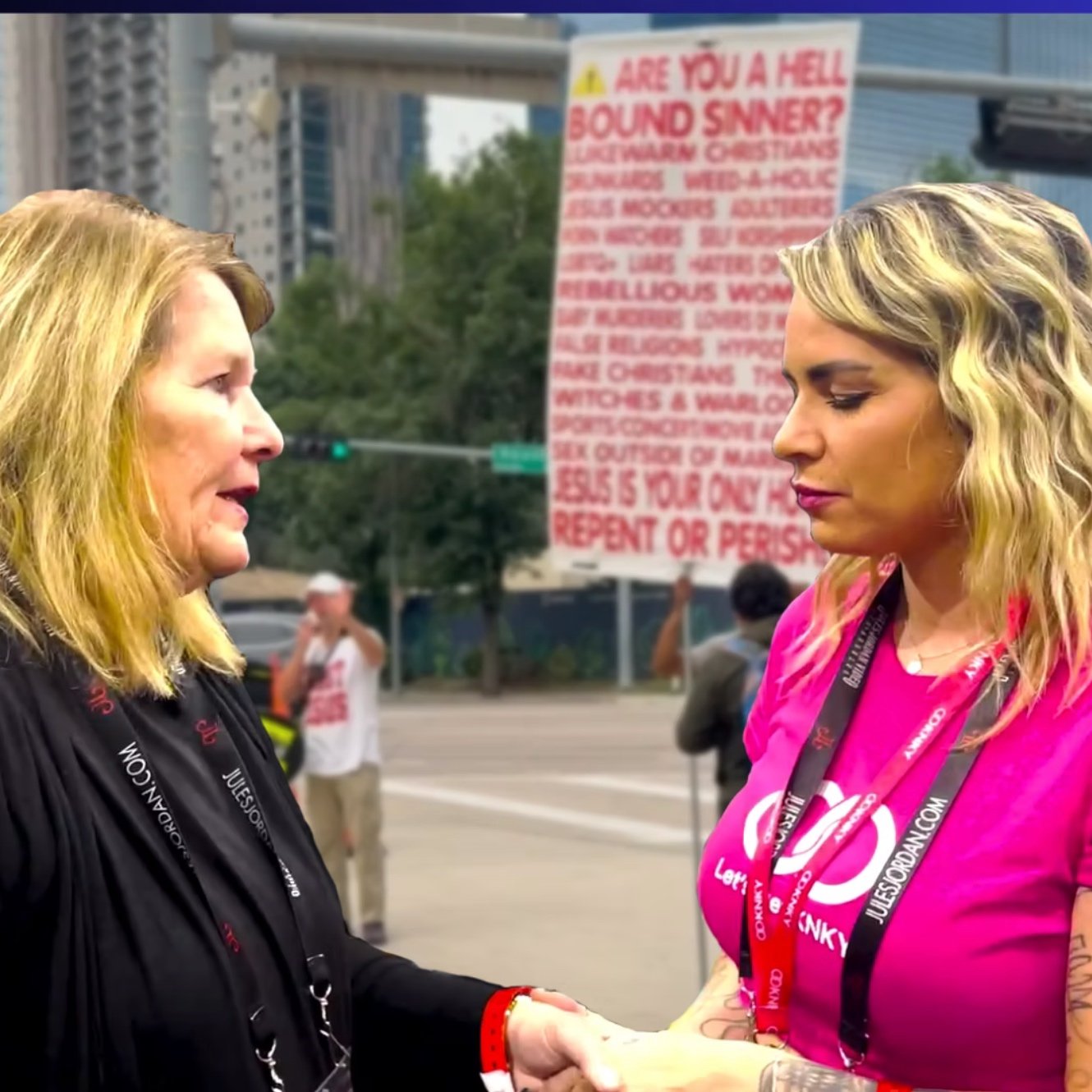 Two women with blonde hair shaking hands at a public event, with a large banner in the background displaying red text on a white background.