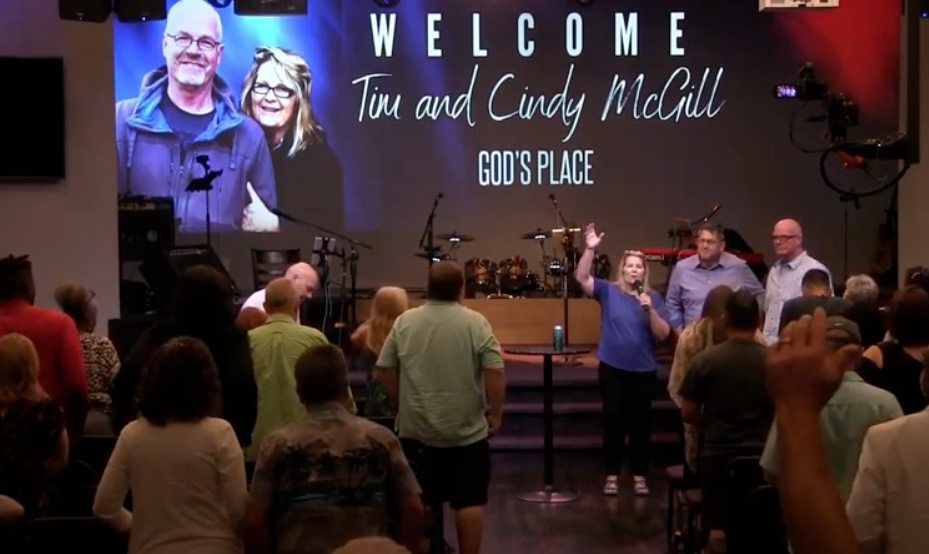 A church service with a large congregation and a stage featuring a large welcome screen displaying the names Tim and Cindy McGill and the words 'God's Place.' Several people on the stage are gathered, with one woman speaking into a microphone and others standing beside her.