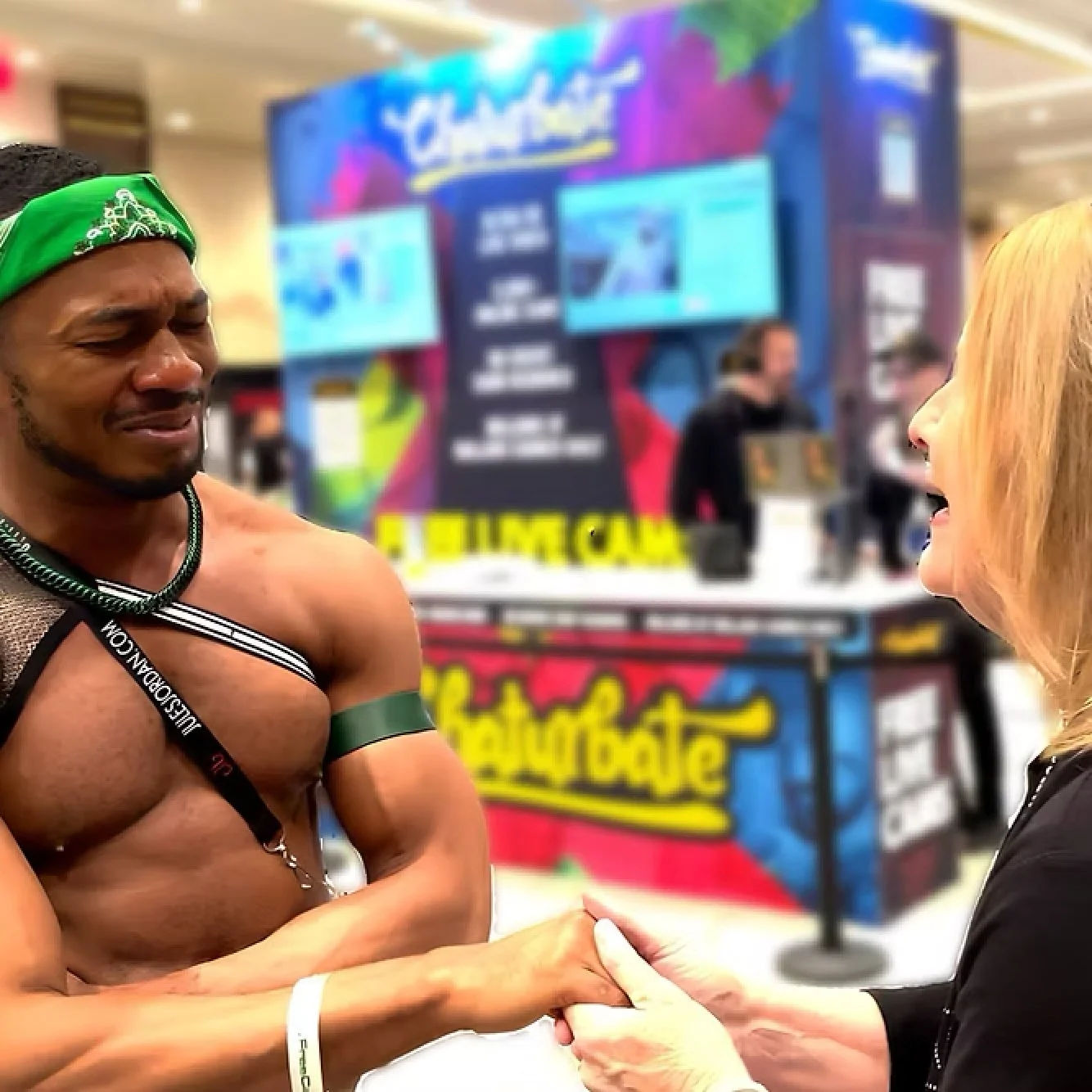 A shirtless man with a green headband and arm band shaking hands with a woman with blonde hair, at a convention or event with colorful banners in the background.