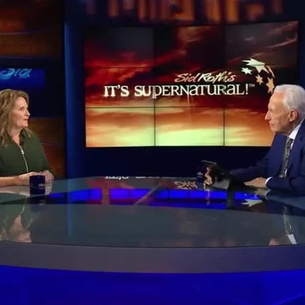 TV studio interview with two people, a woman on the left and a man on the right, during 'Sid Roth's It's Supernatural!' show, with a large screen showing the show's logo and a sunset background in the background.