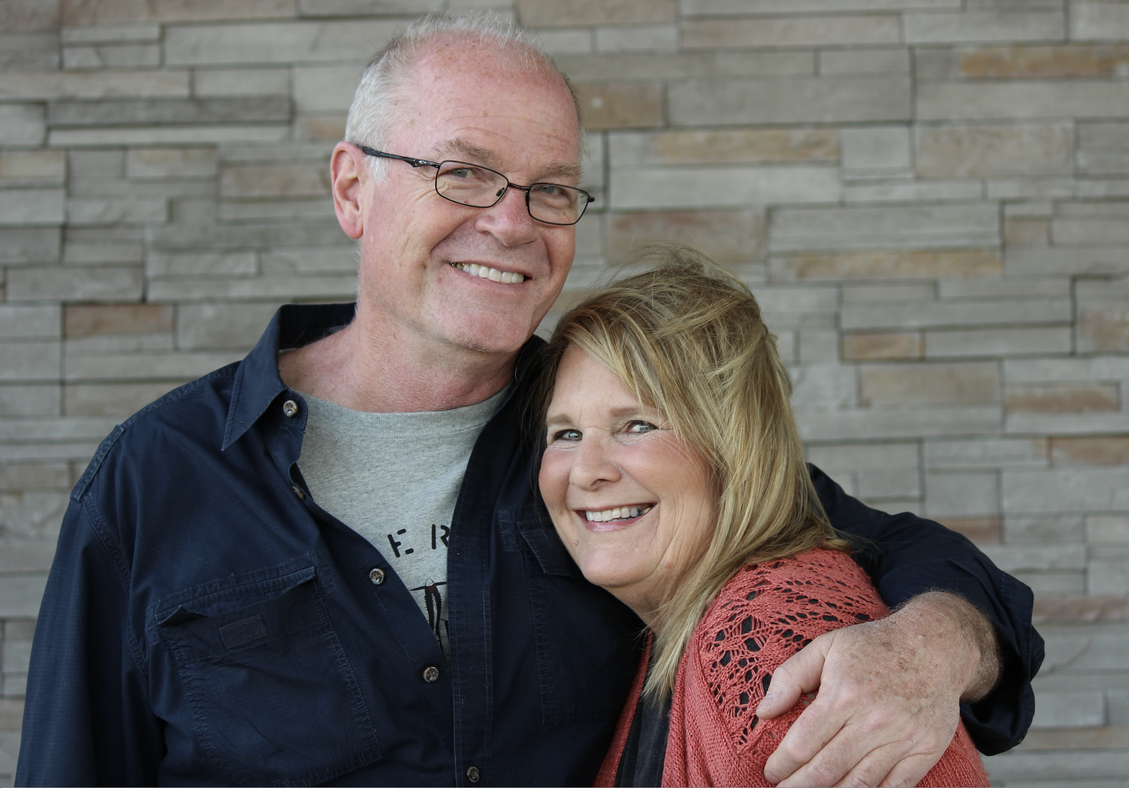 A smiling older man with glasses and a woman with blonde hair embracing in front of a brick wall.