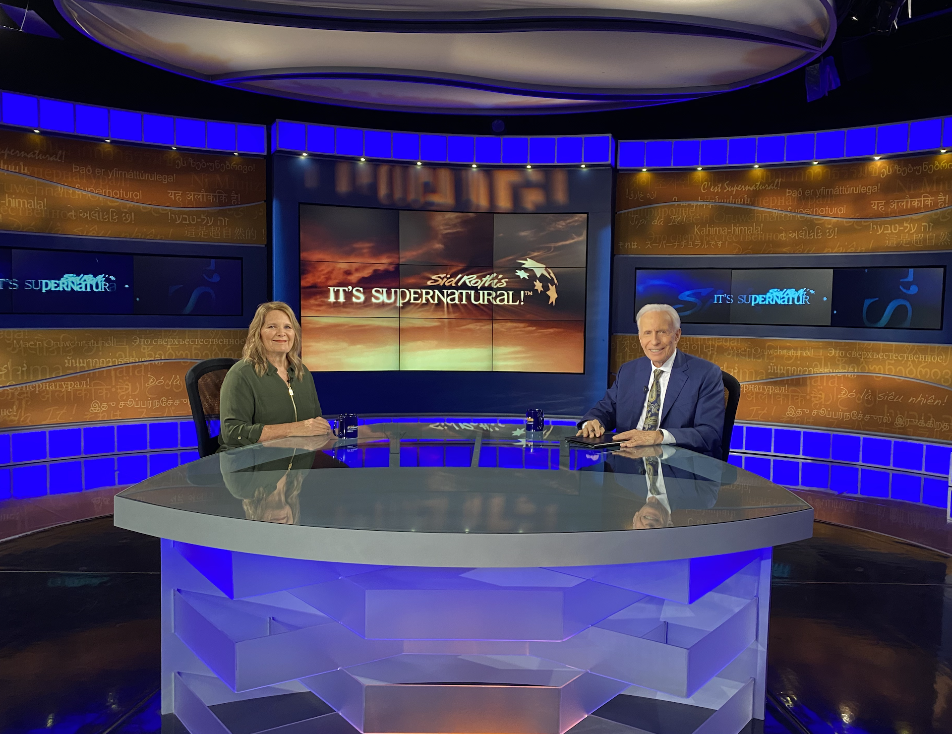 Two people sitting at a glass table in a TV studio with a large screen behind them displaying the text "Sid Roth's It's Supernatural!" The woman is on the left and the man is on the right, both smiling. The studio has vibrant blue and orange lighting.