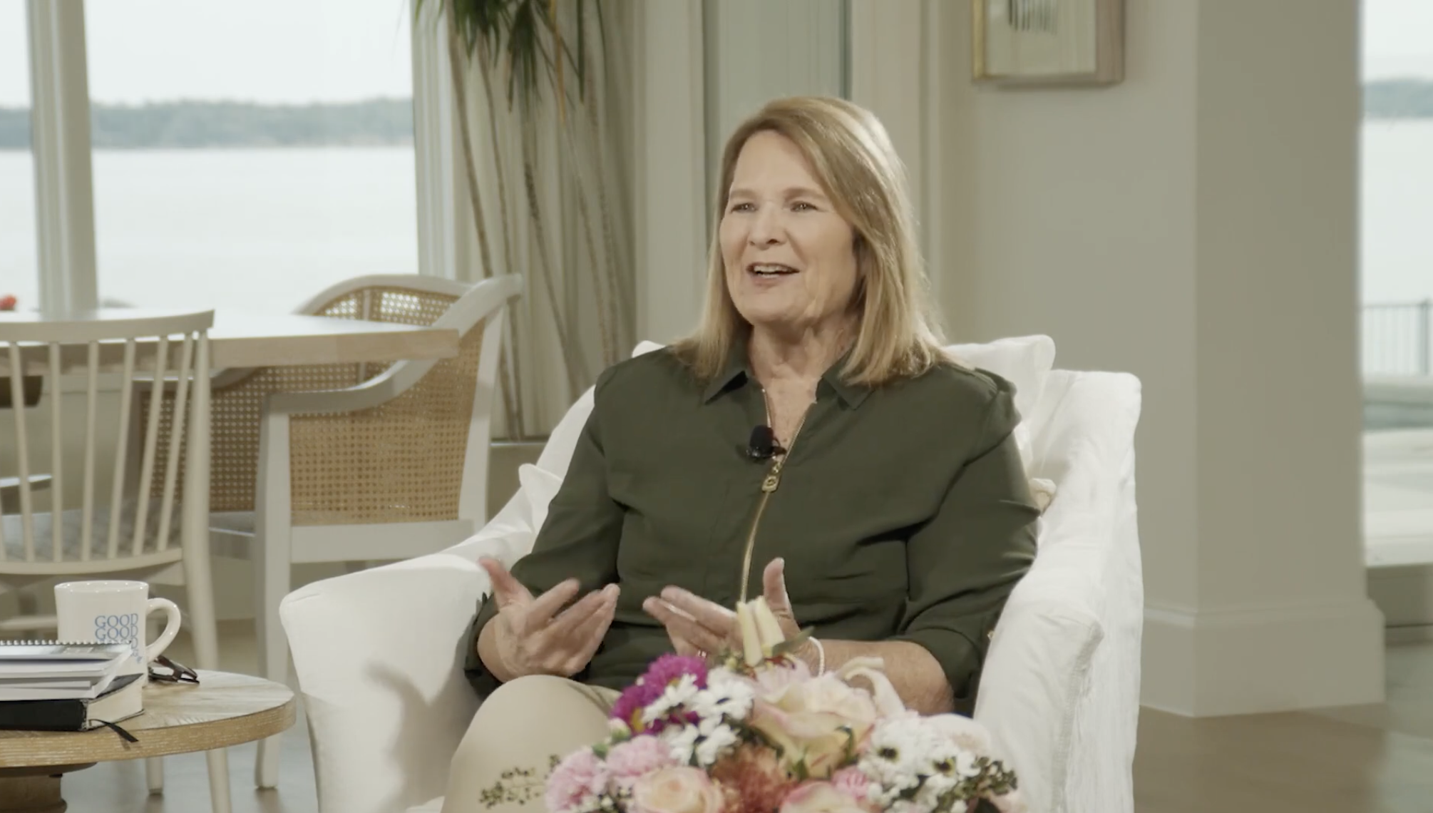 Woman with shoulder-length light brown hair speaking during an interview, sitting on a white chair near a window with a view of a body of water, a bouquet of pink and white flowers on her lap.