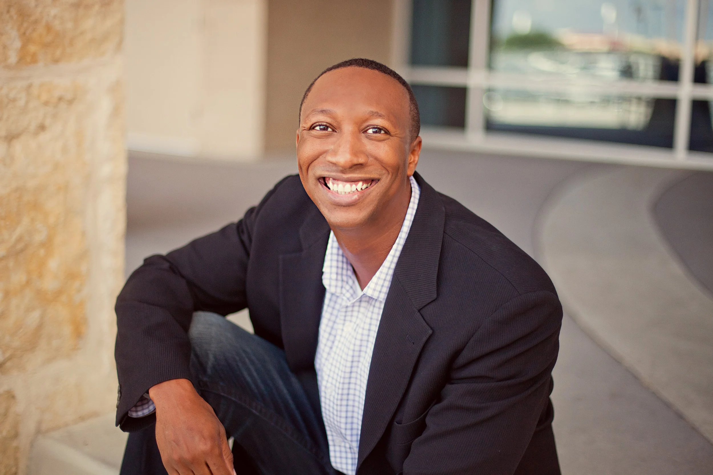 Smiling man dressed in a dark blazer and checkered shirt sitting outdoors near a window and a stone pillar.