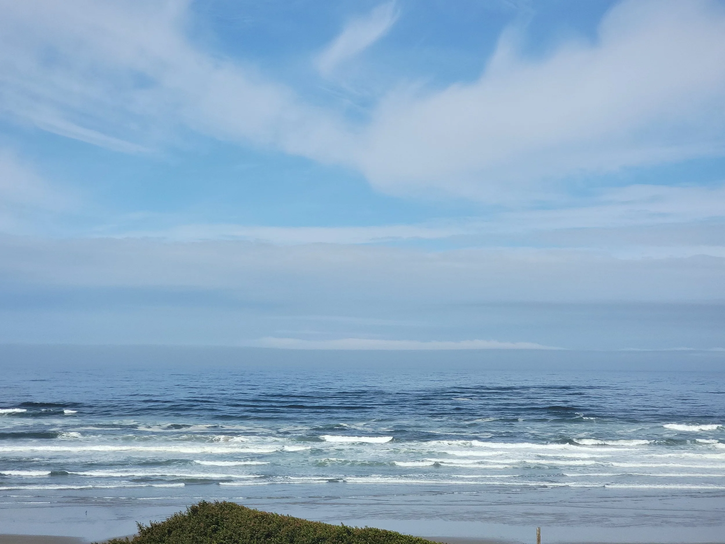 A view of the ocean with waves crashing onto the sandy beach and blue sky with scattered clouds above.