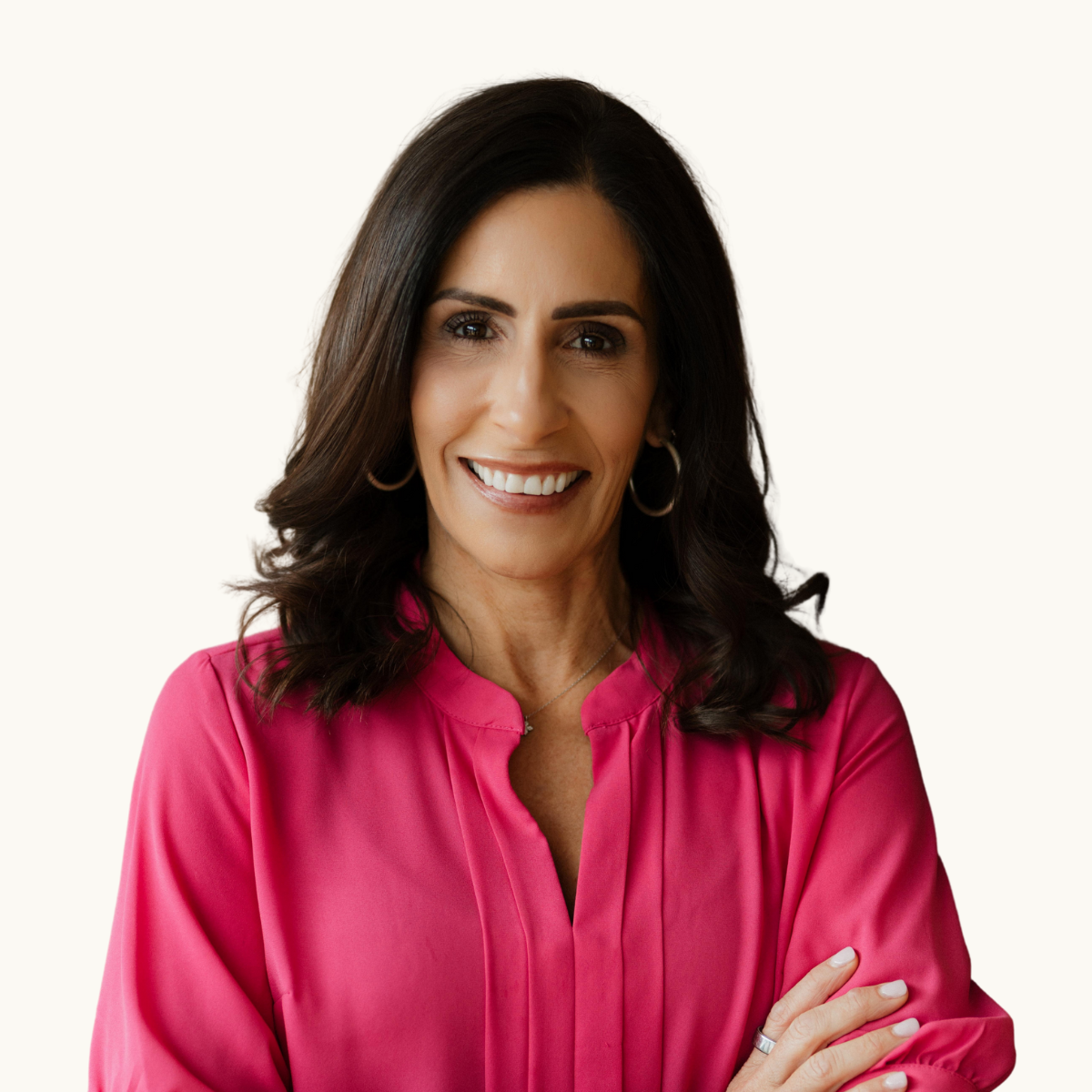 Portrait of a woman with dark brown hair, wearing a pink blouse, smiling with crossed arms against a plain white background.