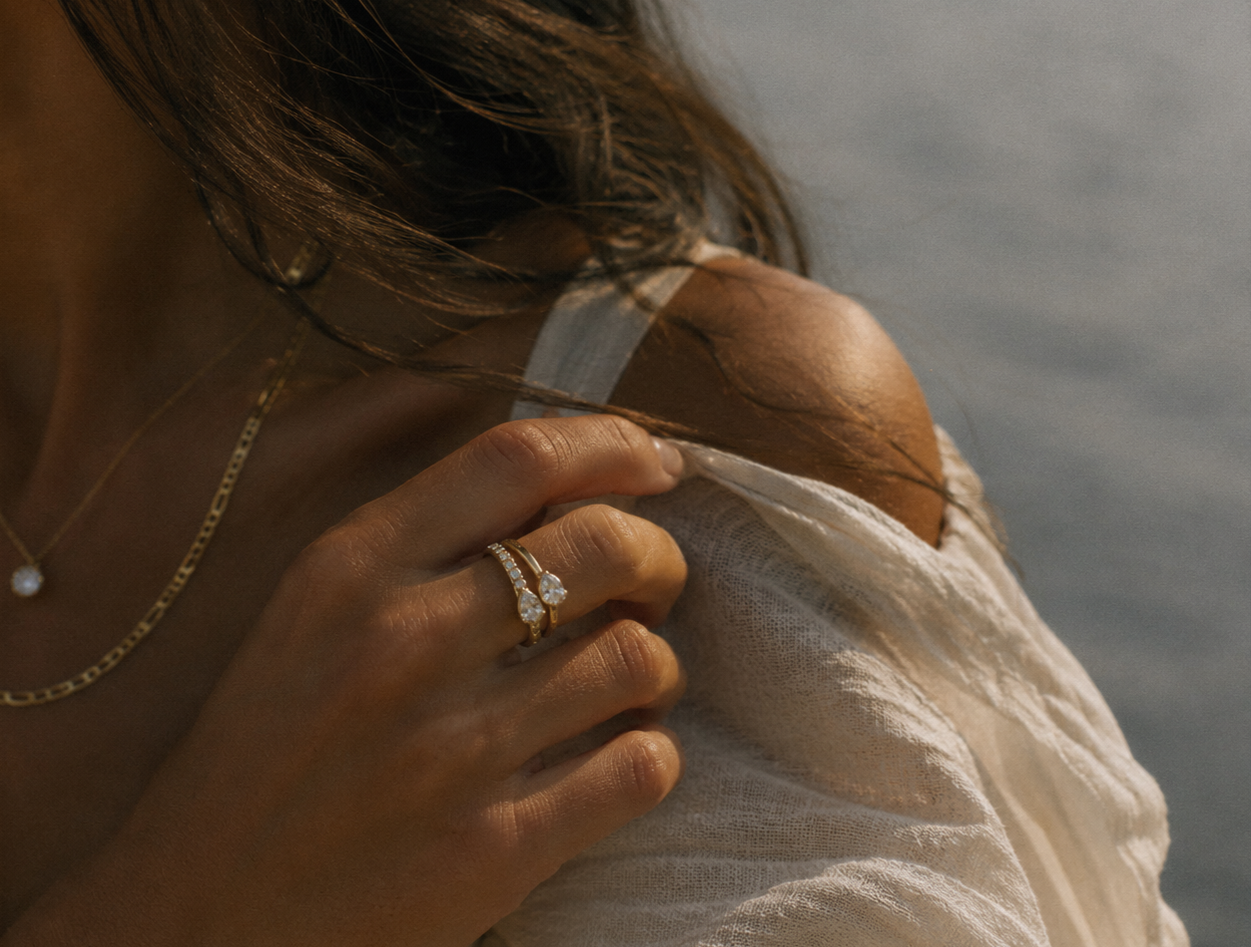 A woman in a white lace top showing her shoulder with jewelry, including a ring with a large gemstone and layered necklaces.