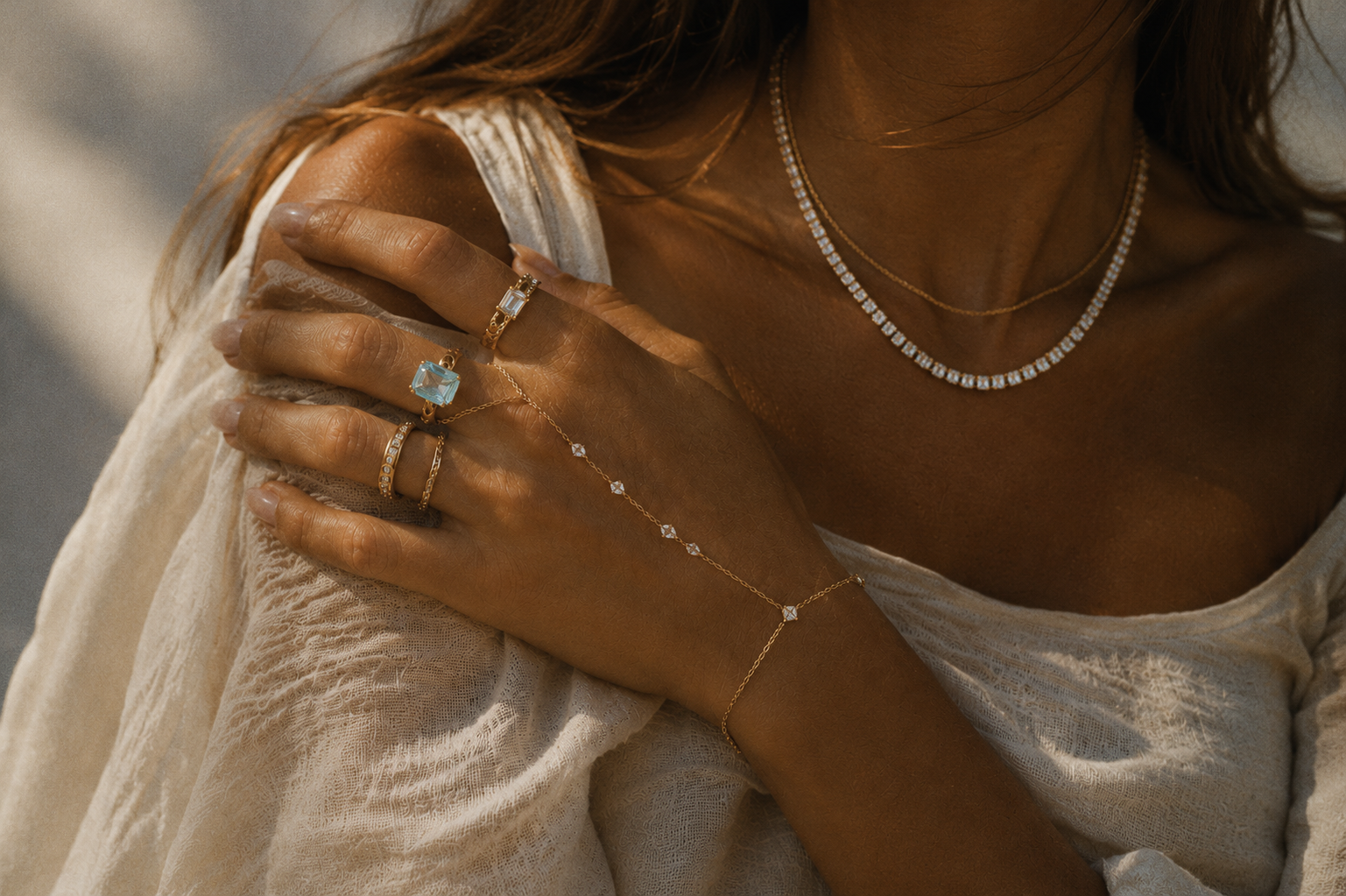 Close-up of a woman wearing gold jewelry, including a diamond choker necklace, a layered chain necklace, several rings with blue gemstones, a bracelet, and a delicate hand chain, with her hand resting on her shoulder in a light-colored, textured fabric top.