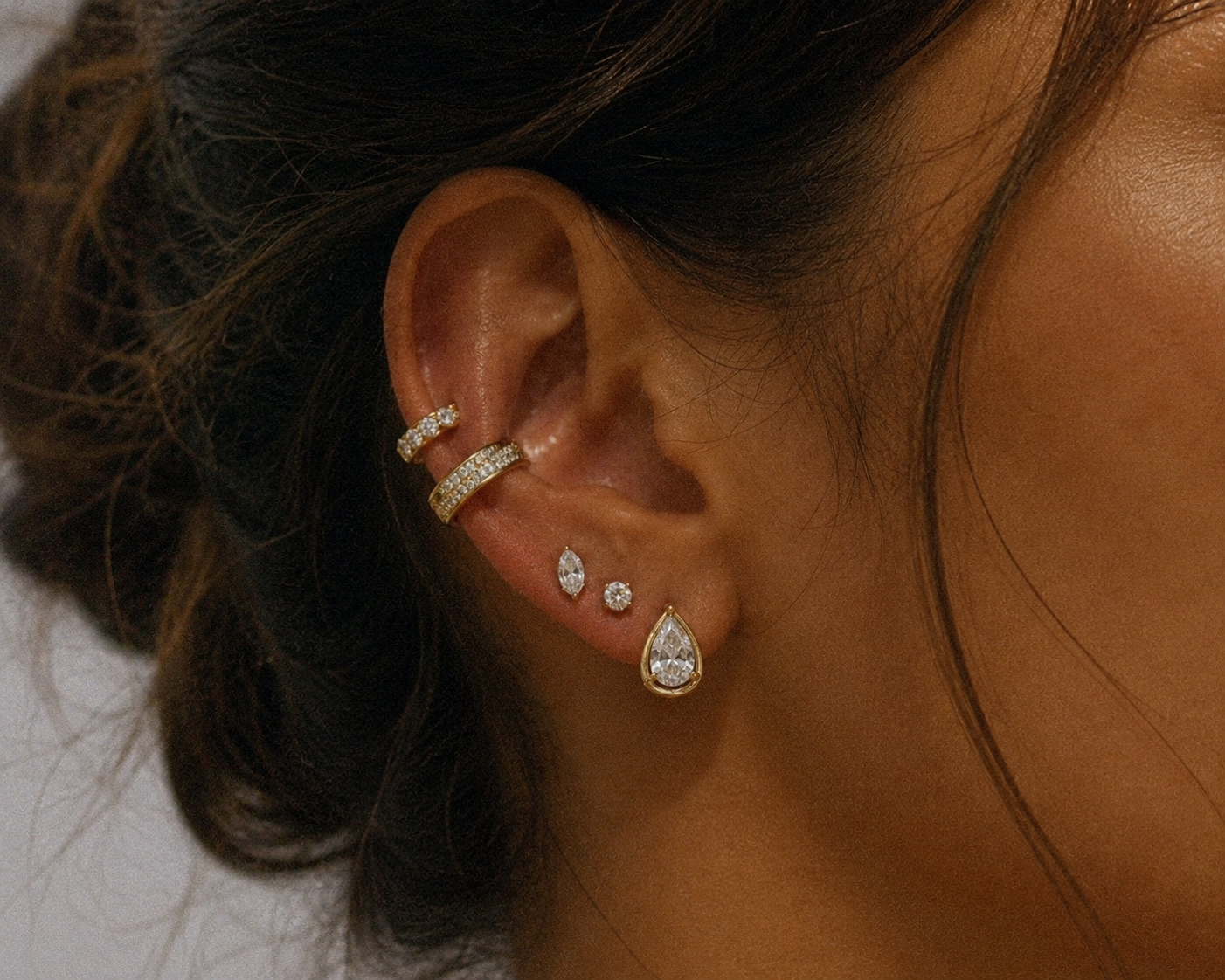 Close-up of a woman's ear with multiple earrings, including a large teardrop-shaped diamond earring and several smaller diamond-studded hoops and studs.