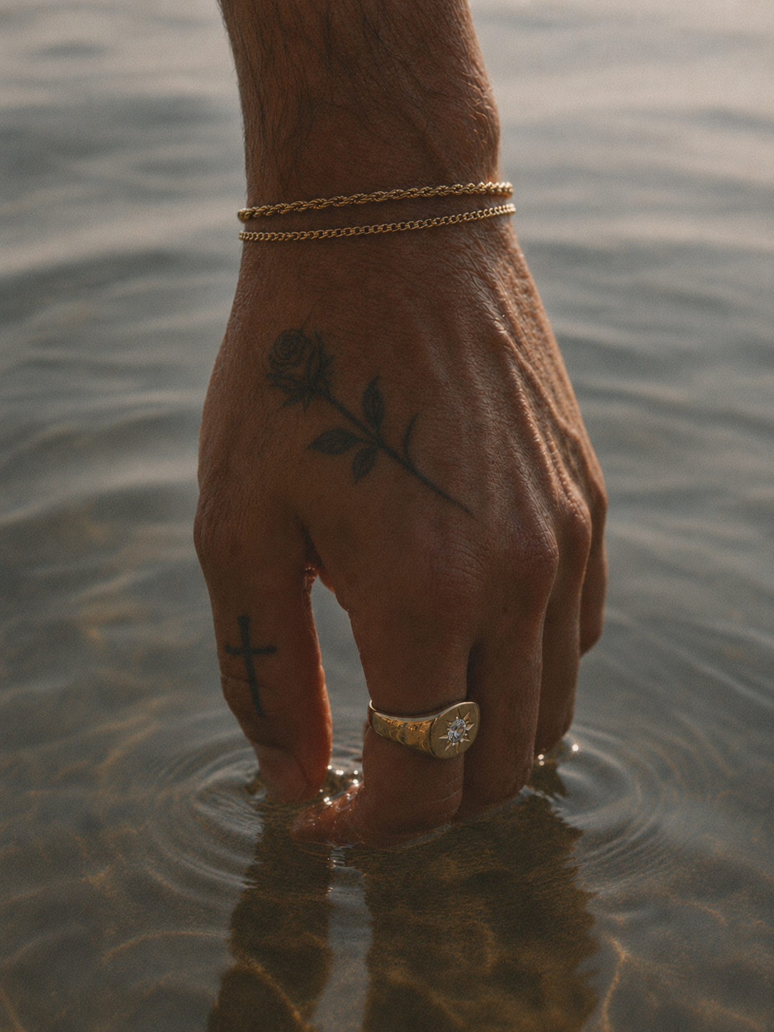Close-up of a person's hand submerged in water, showing tattoos of a cross and a flower, wearing gold rings and bracelets.