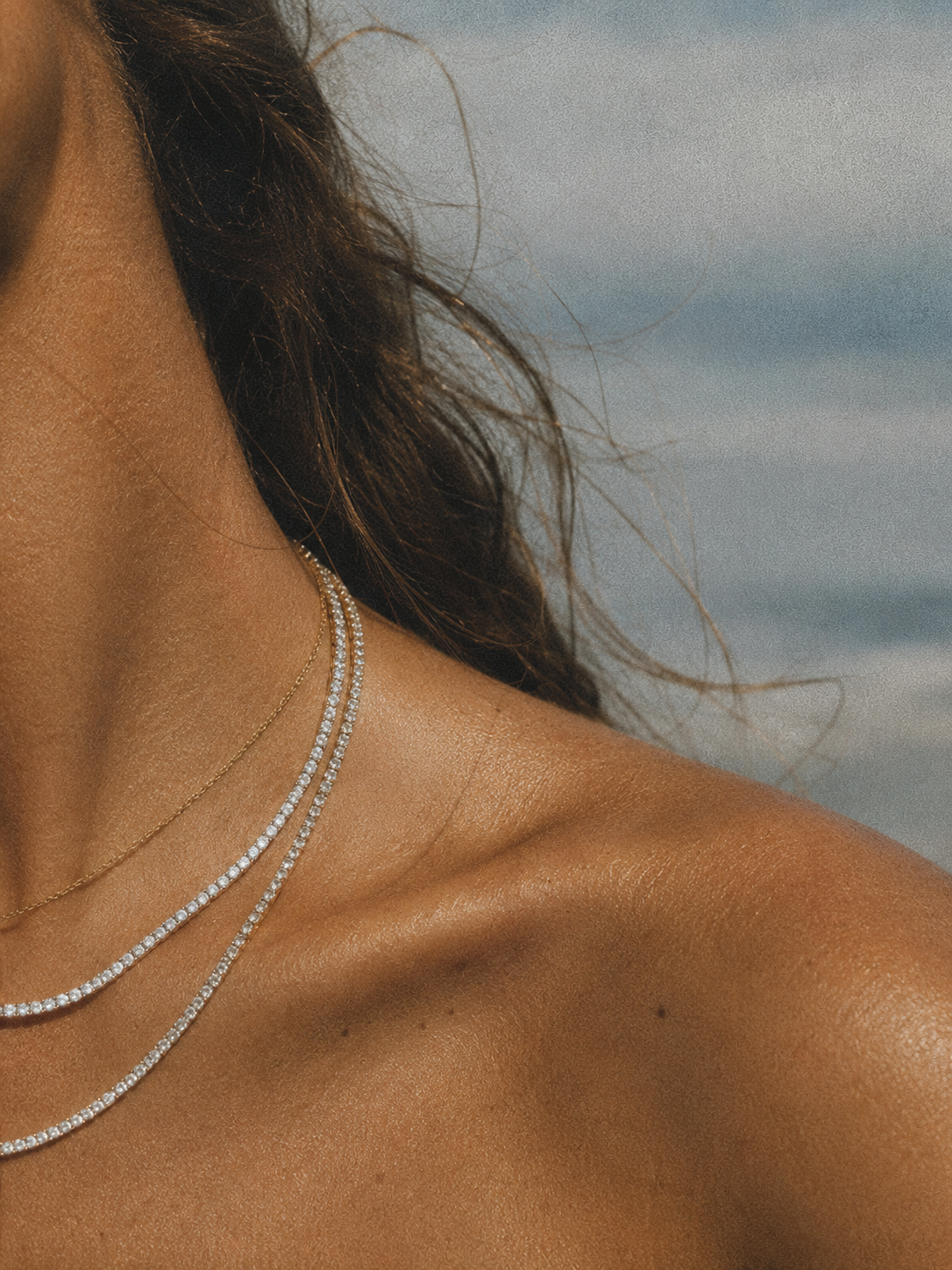 Close-up of a woman's shoulder and neck, with layered diamond necklaces and wavy brown hair, against a blurred cloudy sky background.
