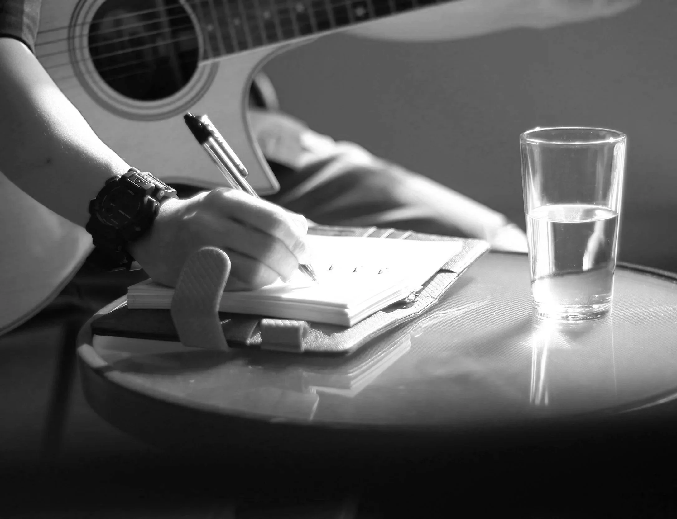 A person with a watch on their wrist is writing in a notebook with a pen. There is a glass of water on the table and a guitar in the background.