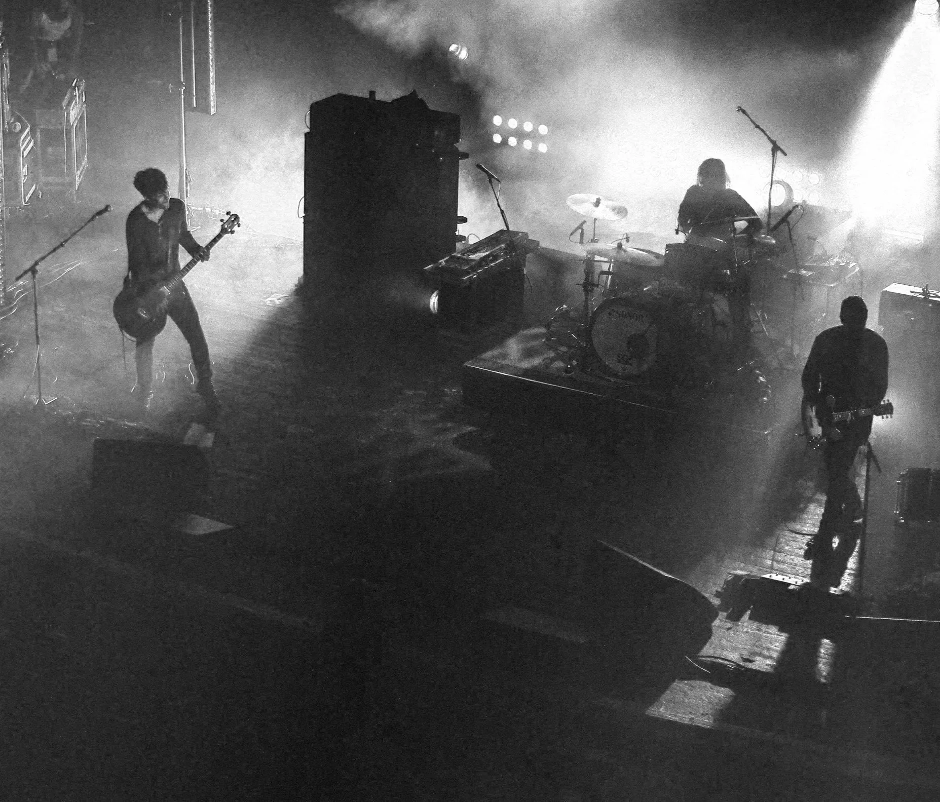 Black and white photograph of a band performing on stage with three musicians playing guitars and a drummer behind a drum set, stage lights creating a smoky atmosphere.