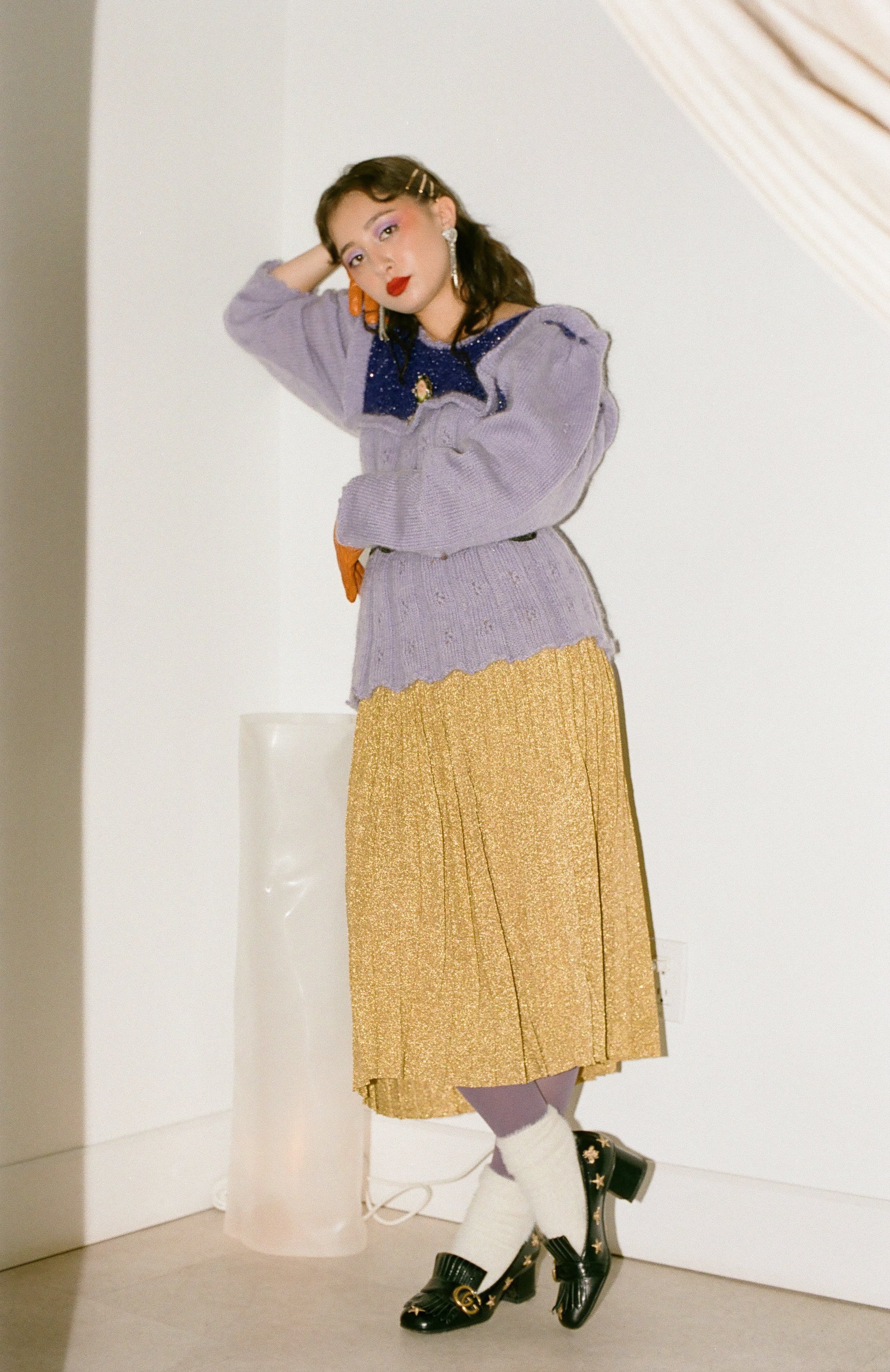 Femme portant un pull violet, une jupe longue dorée, des chaussettes blanches et des bottines noires avec des motifs. Elle pose de profil, avec la main derrière la tête dans une pièce lumineuse et nue.