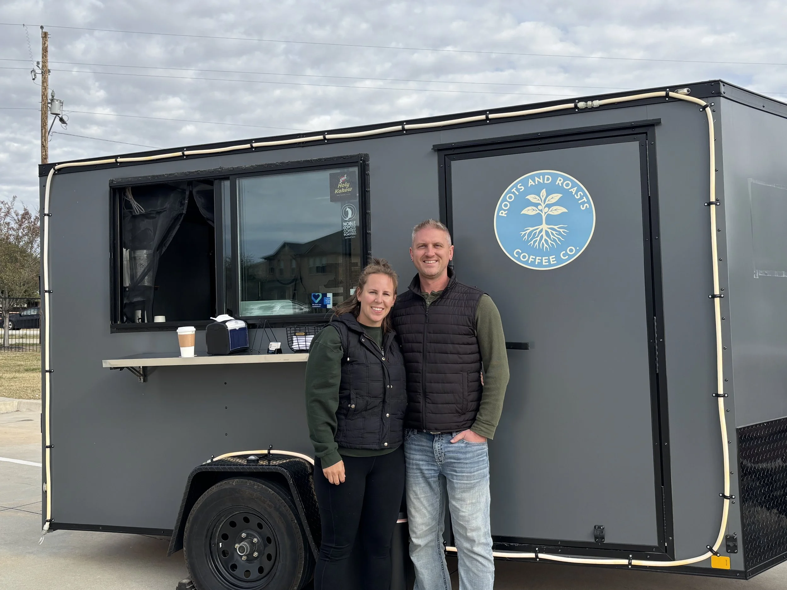 Two people standing in front of a dark gray coffee trailer with the logo 'Roots and Roasts Coffee Co.' visible on the side.