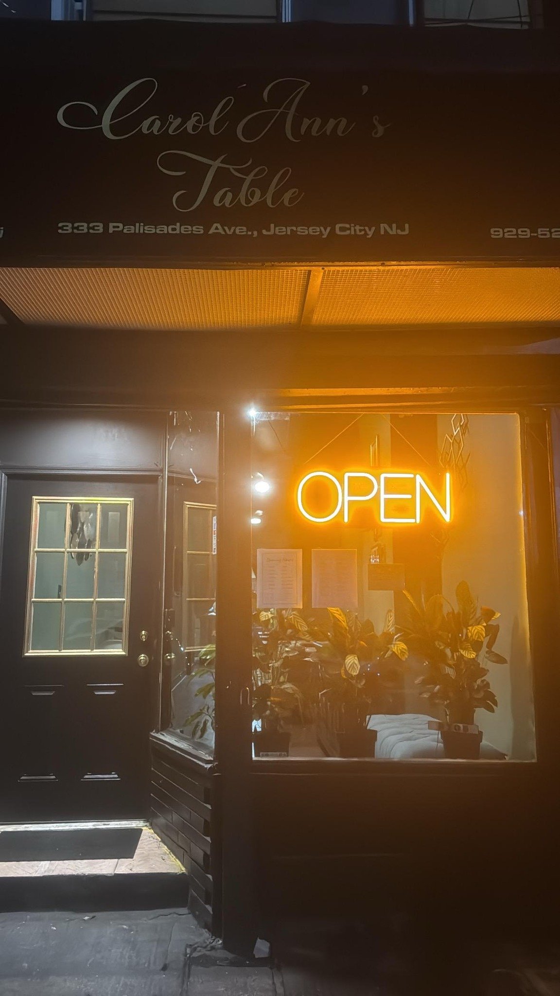The entrance of a restaurant or cafe with a neon 'OPEN' sign in the window, potted plants inside, and a sign above with the name 'Carol Ann's Table' and the address '333 Palisades Ave., Jersey City NJ'.