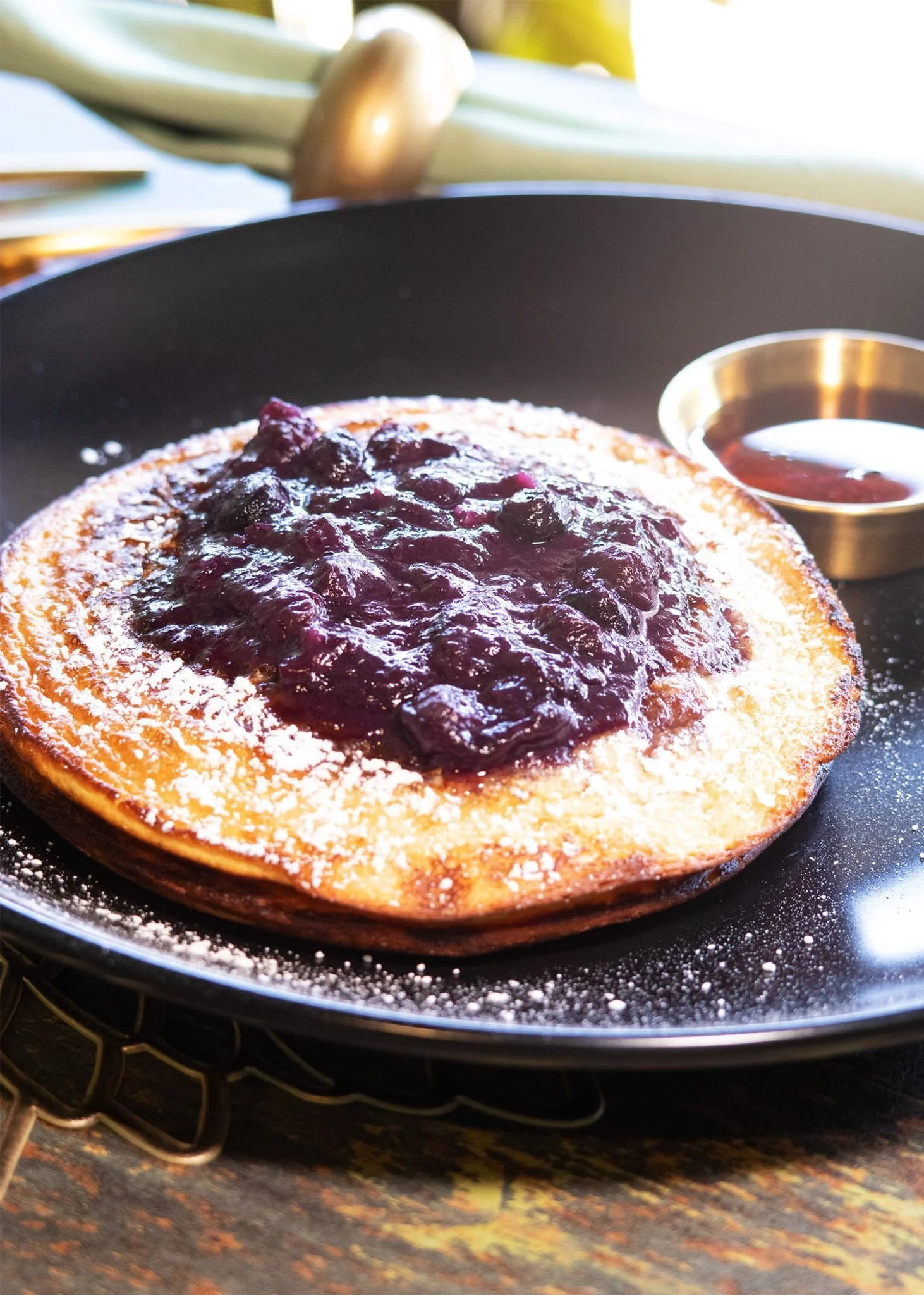 A golden-brown pancake topped with blueberry compote, powdered sugar, served on a black plate with a small dish of syrup.
