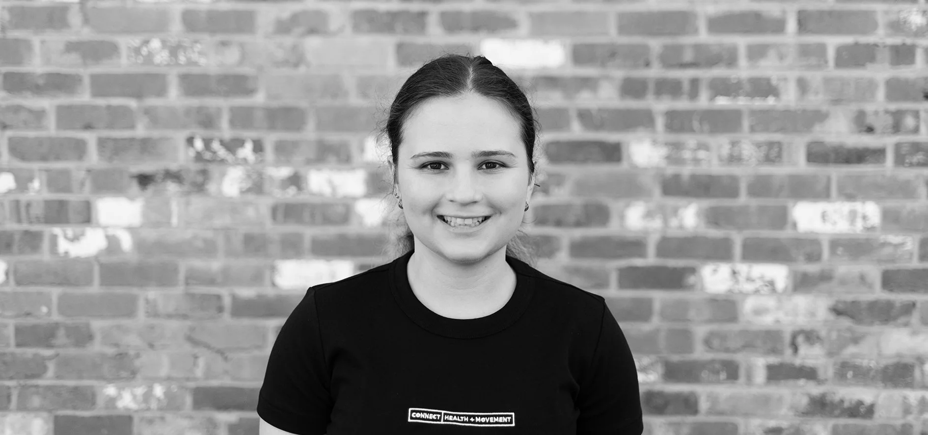 Young woman smiling, wearing a black shirt with logo, standing in front of brick wall