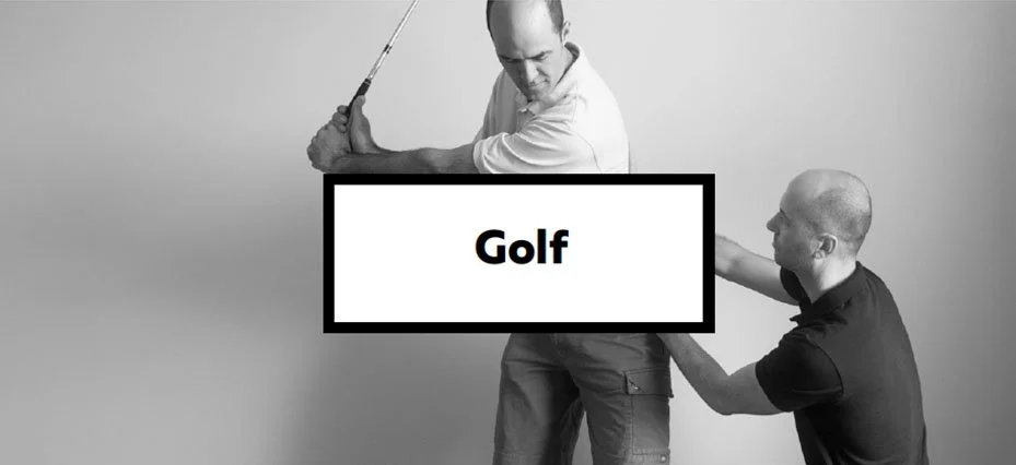 Two men practicing golf in a studio, one holding a golf club preparing to swing, the other assisting or coaching, with a large sign in the center reading 'Golf'.