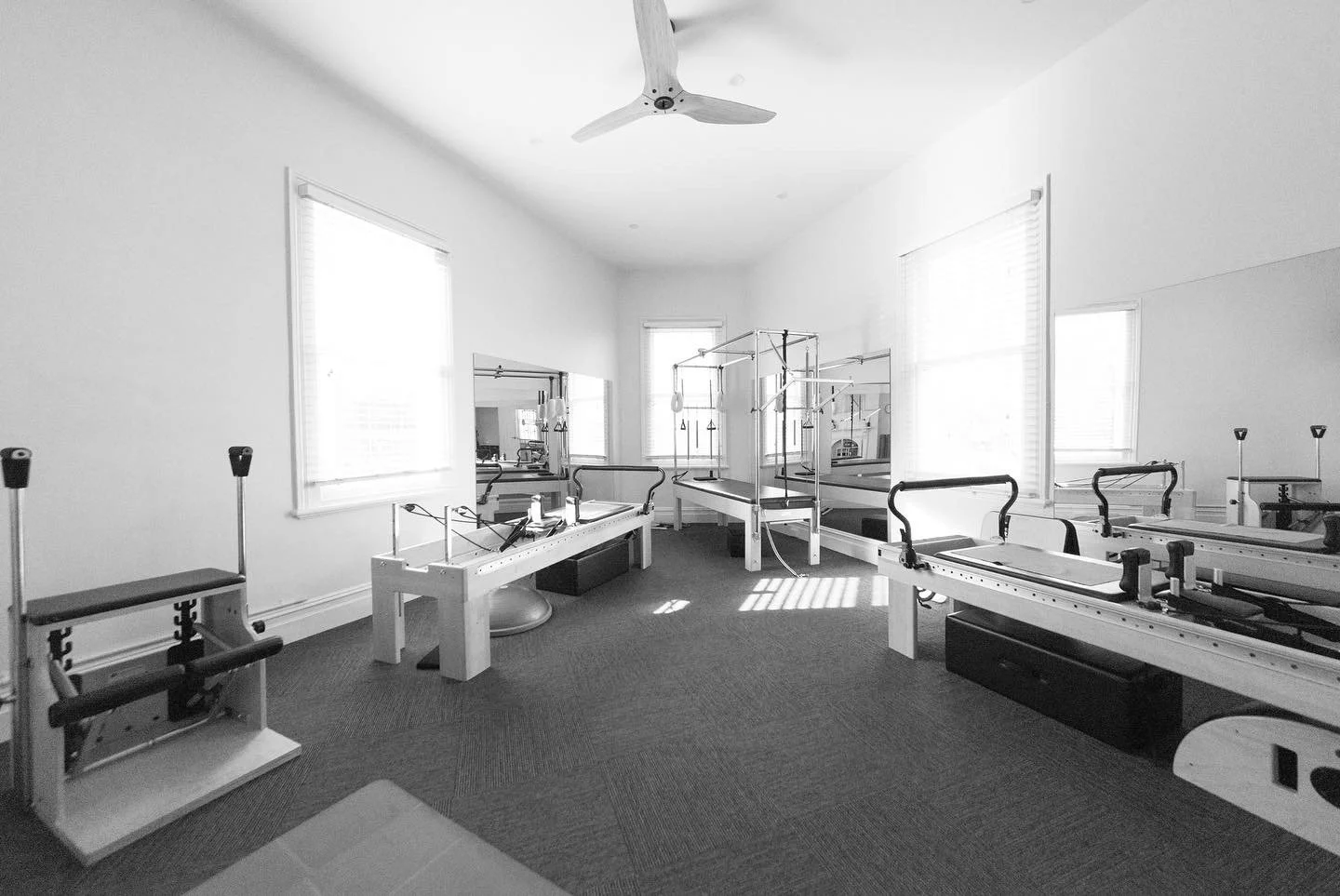A physical therapy or rehabilitation room with Pilates reformer machines and exercise equipment, illuminated by natural light from multiple windows.