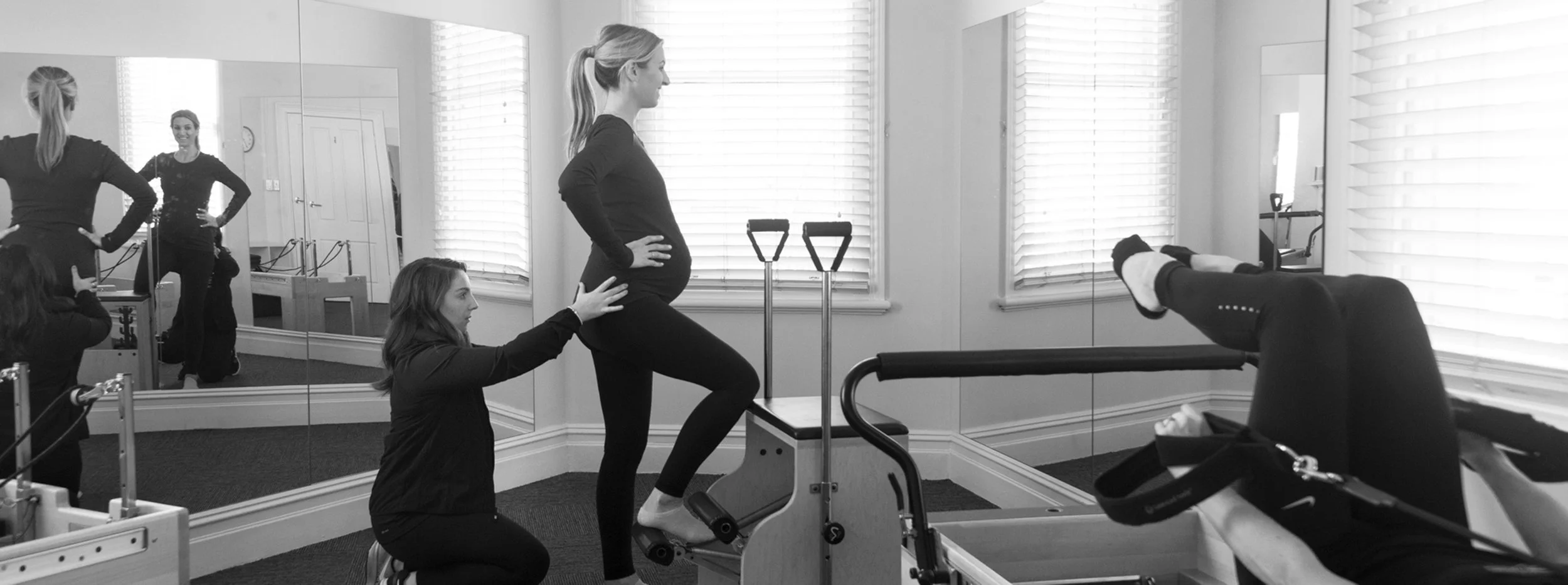 Pregnant woman doing prenatal exercise on Pilates reformer with support from trainer in studio.