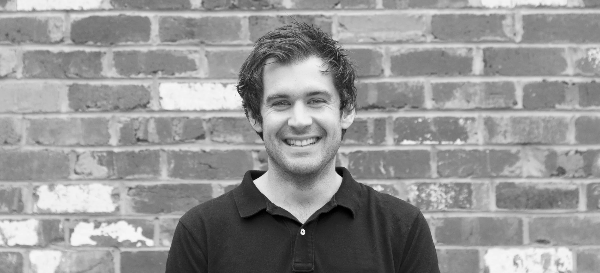 A young man with wavy dark hair and a big smile standing in front of a brick wall, wearing a black polo shirt.