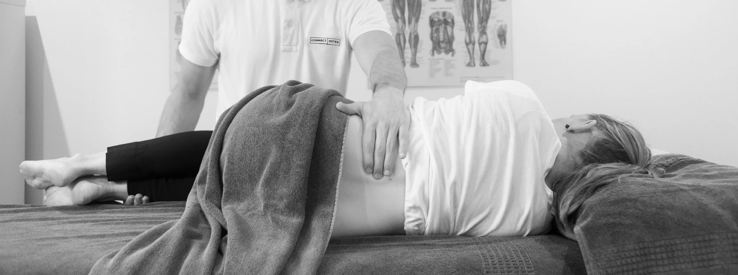 A healthcare professional performing a physical examination on a woman lying on a medical table, with a human anatomy poster on the wall in the background.