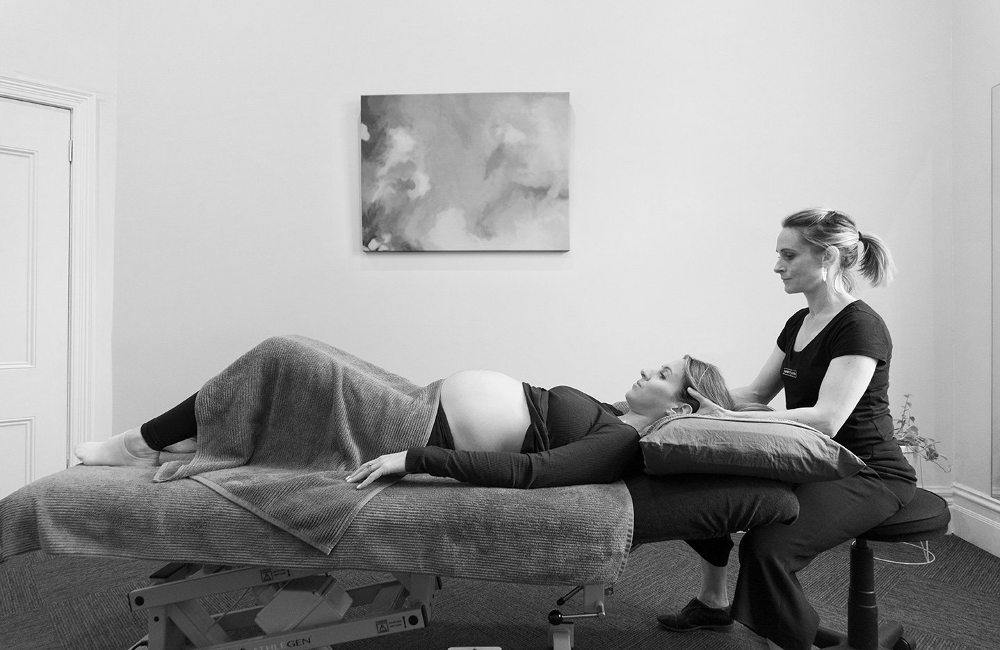 A woman lying on a massage table while a therapist provides a treatment or consultation in a clinical setting.