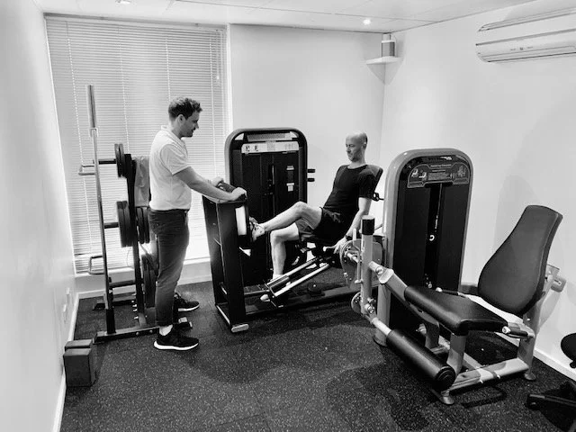A woman exercising on a leg press machine while a man assists her in a home gym with weight equipment and a window with blinds in the background.