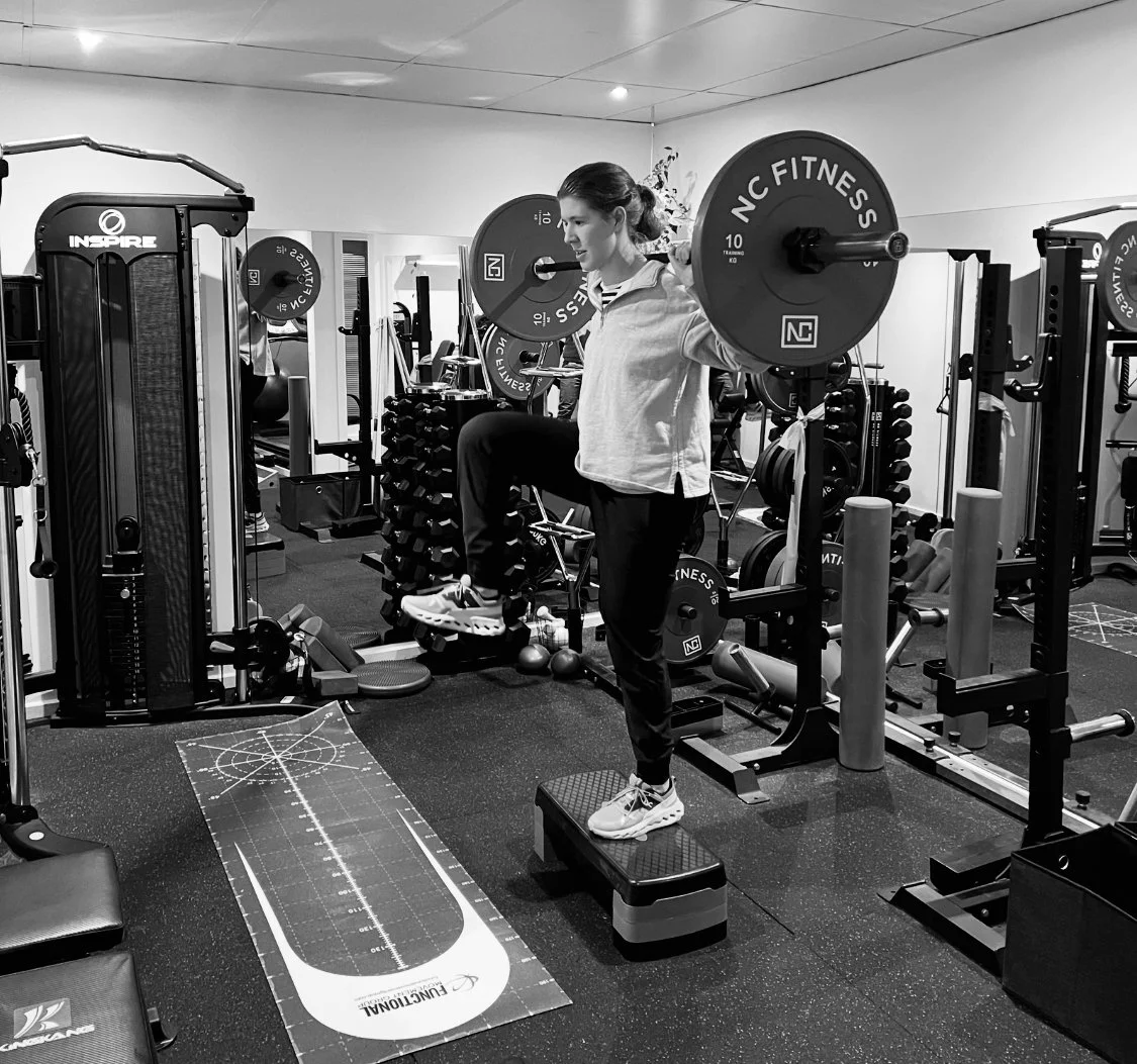 A woman in workout clothes standing on a step platform in a gym, lifting a barbell with weights on her shoulders.