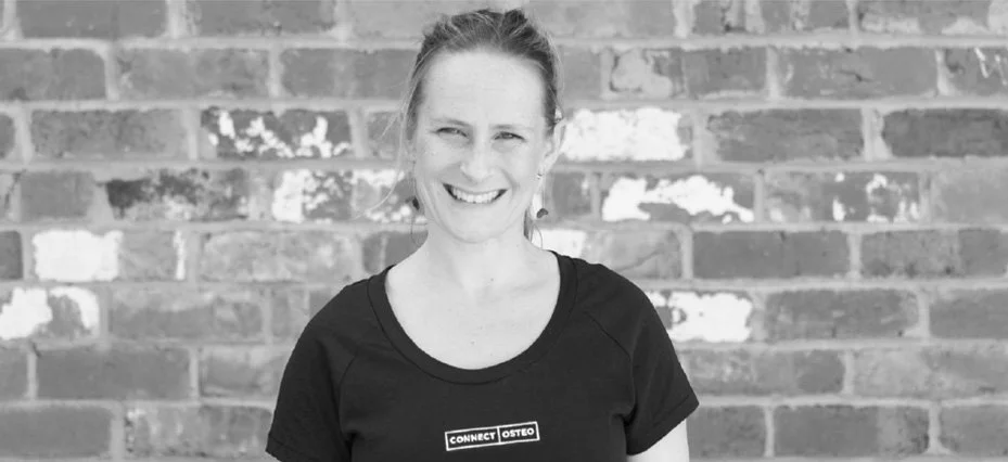 A woman smiling, standing in front of a brick wall, wearing a black t-shirt with a logo that reads 'CONNECT OSTEOPATHY'.