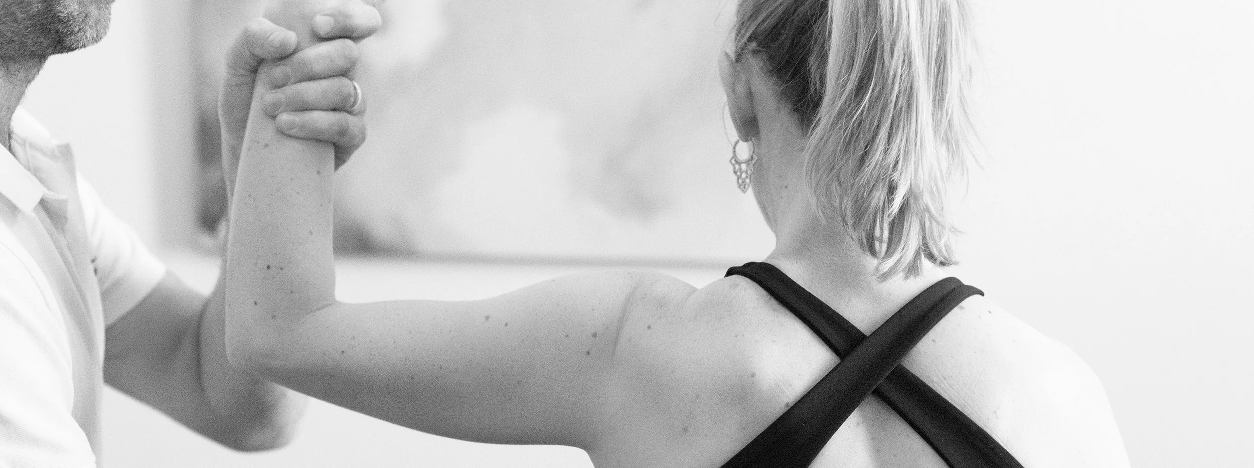 A woman in a sports bra and earrings is arm wrestling with a man, whose face is partially visible, in a gym setting.