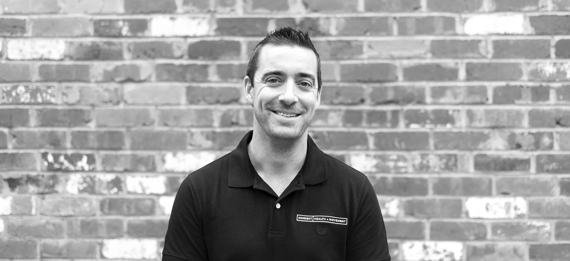 A smiling man with short dark hair wearing a black collared shirt with a logo that reads 'Connect Health + Movement,' standing in front of a brick wall.