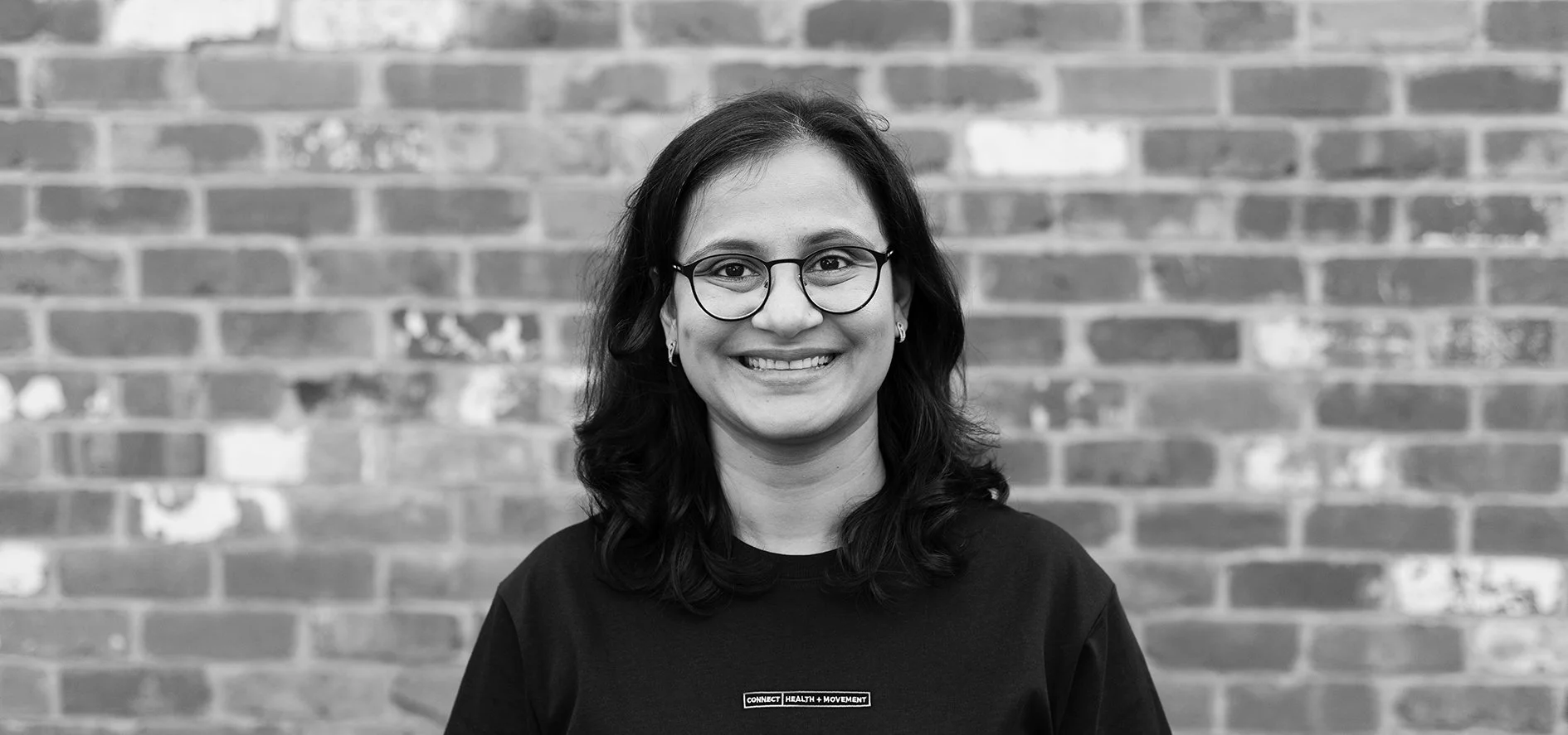 A woman with glasses and earrings, smiling, standing in front of a brick wall.
