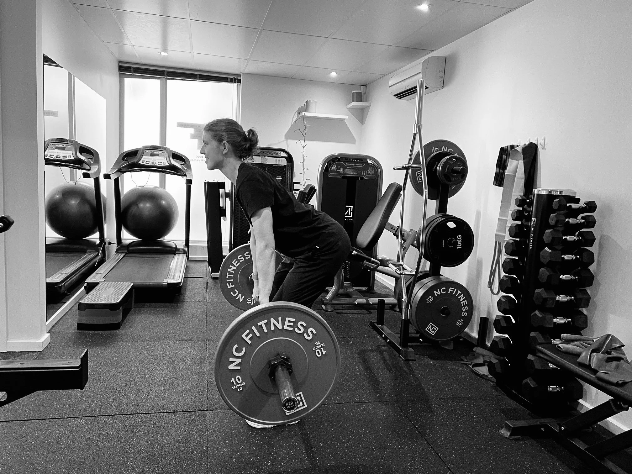 A woman lifting a barbell in a gym with workout equipment including exercise balls, a treadmill, weight plates, and a rack of dumbbells.