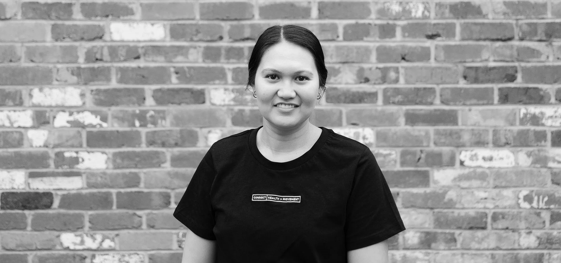 Black and white photo of a woman with dark hair standing in front of a brick wall, wearing a black t-shirt with text that reads 'CONNECT HEALTH + MOVEMENT'.