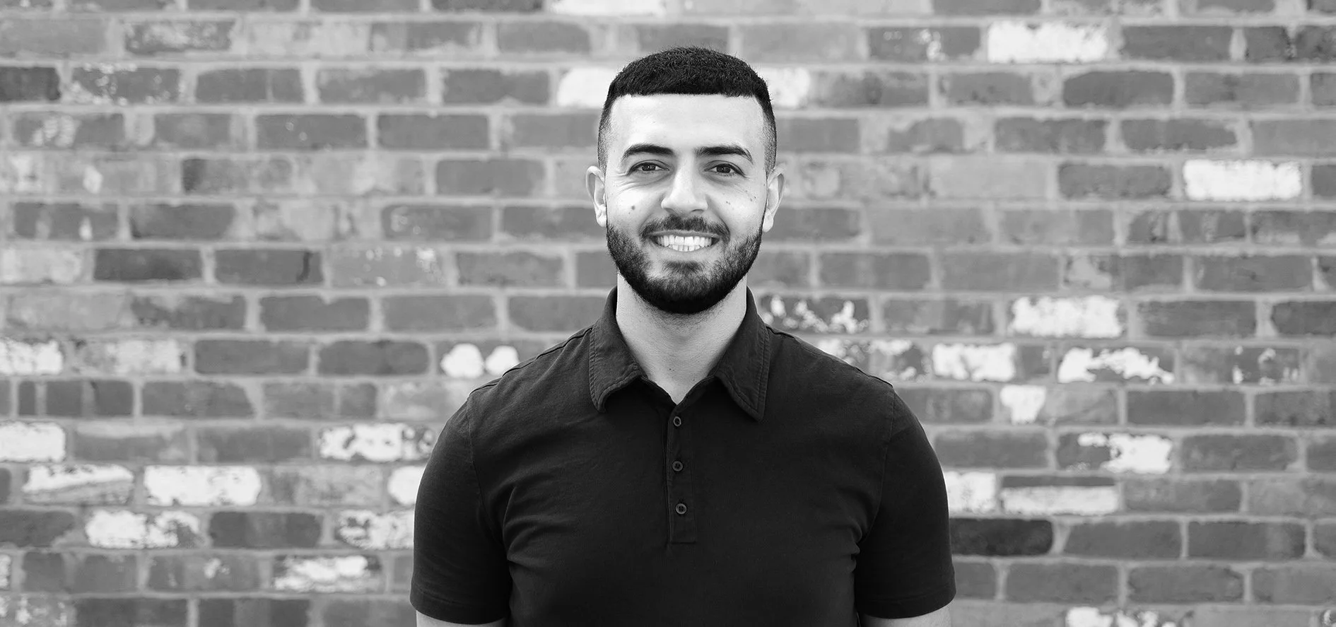 Black and white photo of a smiling man with short dark hair and a beard, wearing a dark button-up shirt, standing in front of a brick wall.