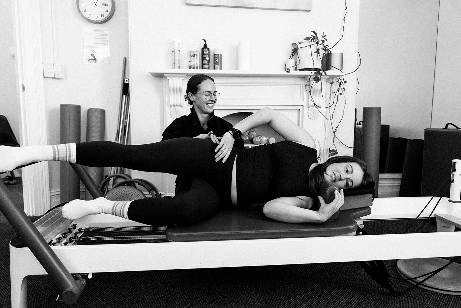 A woman in athletic clothing is pregnant and practicing a fitness exercise on a piece of equipment with support from a trainer in a gym with large windows and mirrors.
