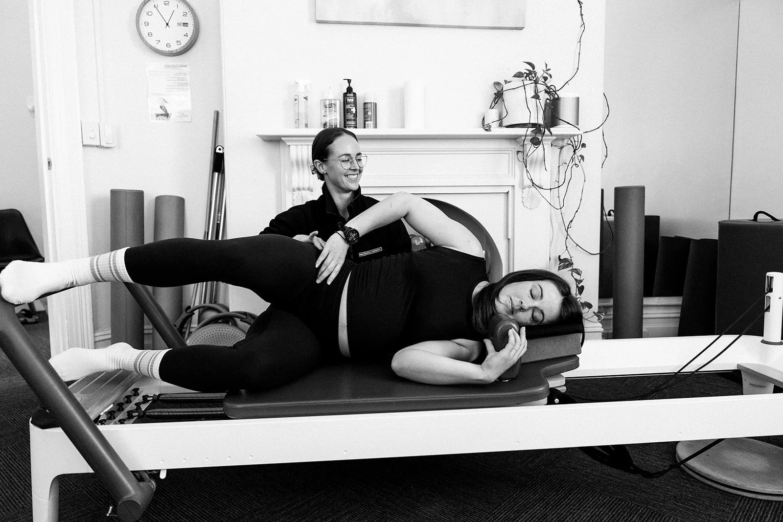 Pregnant woman doing prenatal exercise on Pilates reformer with support from trainer in studio.