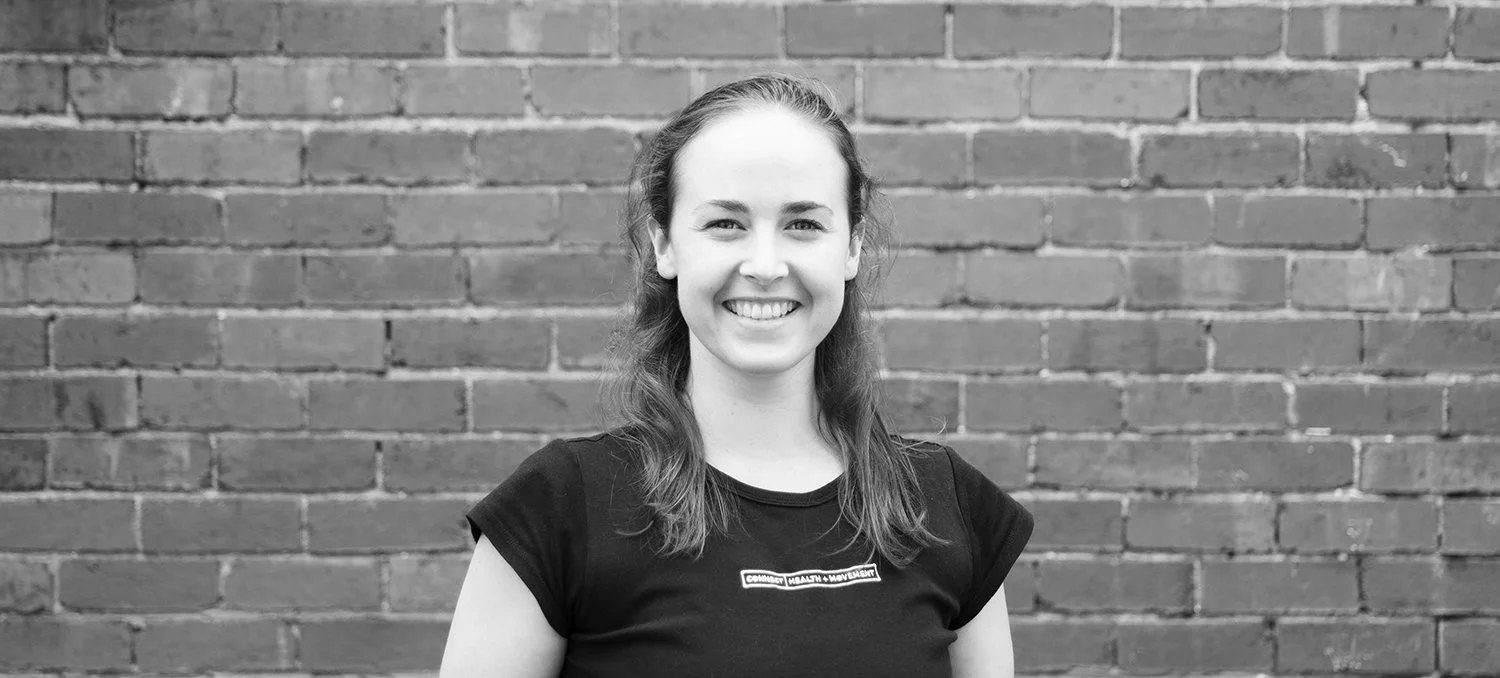 Black and white photo of a smiling woman with long hair standing in front of a brick wall, wearing a black T-shirt.