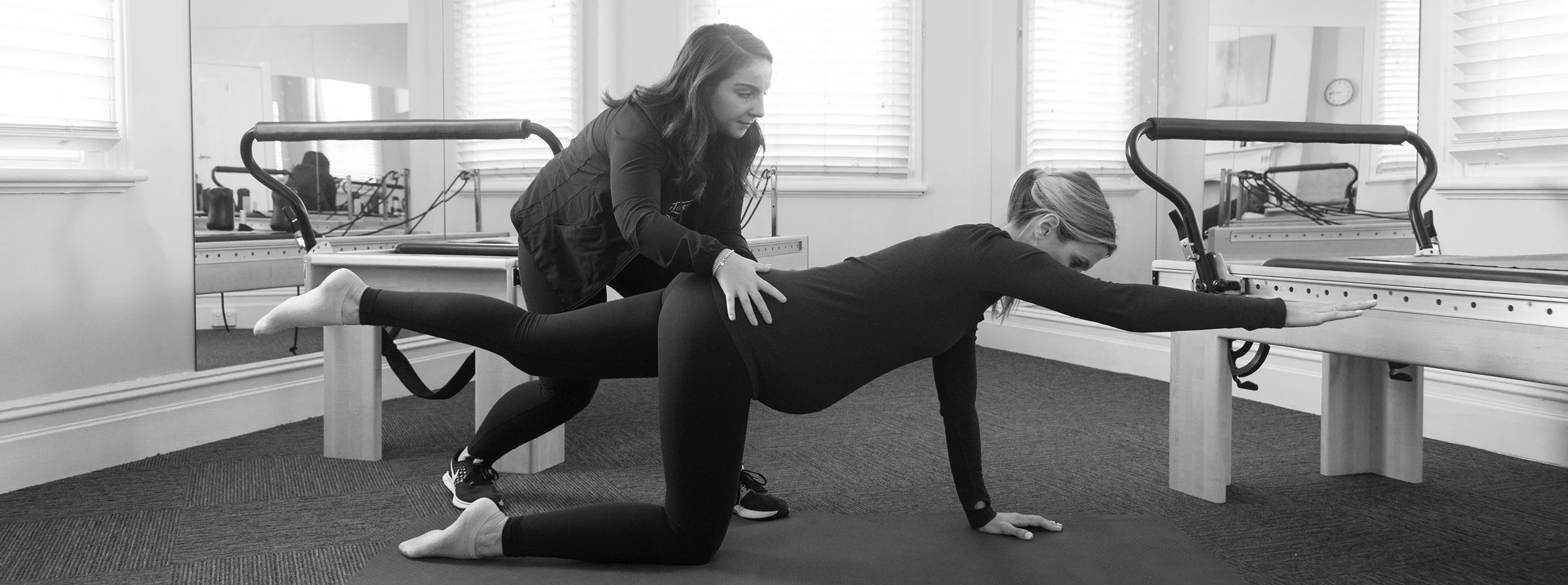 A woman is practicing a yoga pose on a mat while another woman assists her by placing her hands on her back for support in a fitness studio.