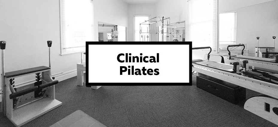 A Pilates studio with various exercise equipment including reformers and a Cadillac in a bright room with large windows, and a sign in the center that says 'Clinical Pilates'.
