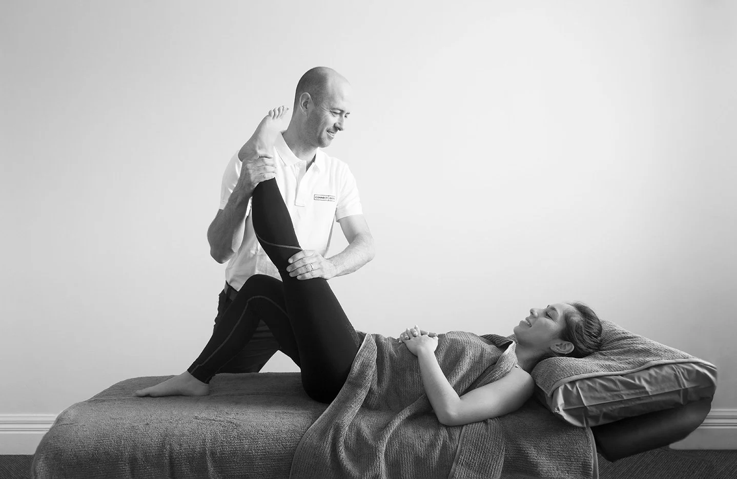 A man giving physical therapy to a woman lying on a bed, holding her leg up during a session.