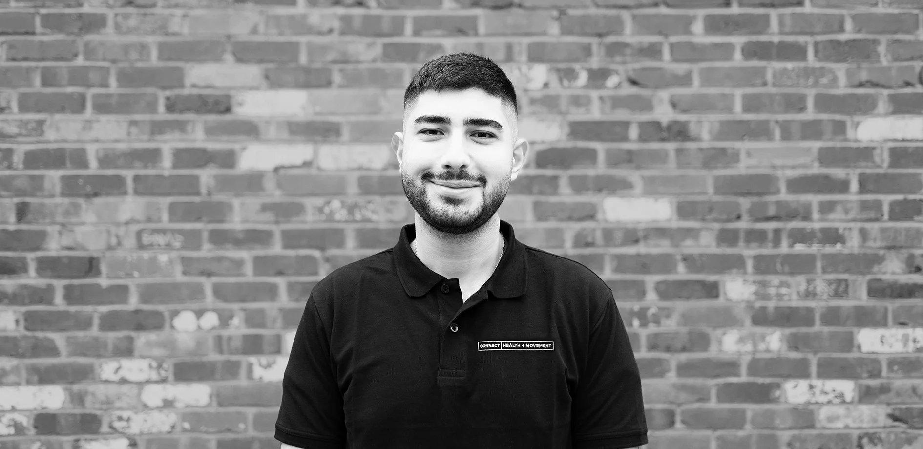 A young man with short dark hair and a beard, wearing a black polo shirt, standing in front of a brick wall, smiling at the camera.