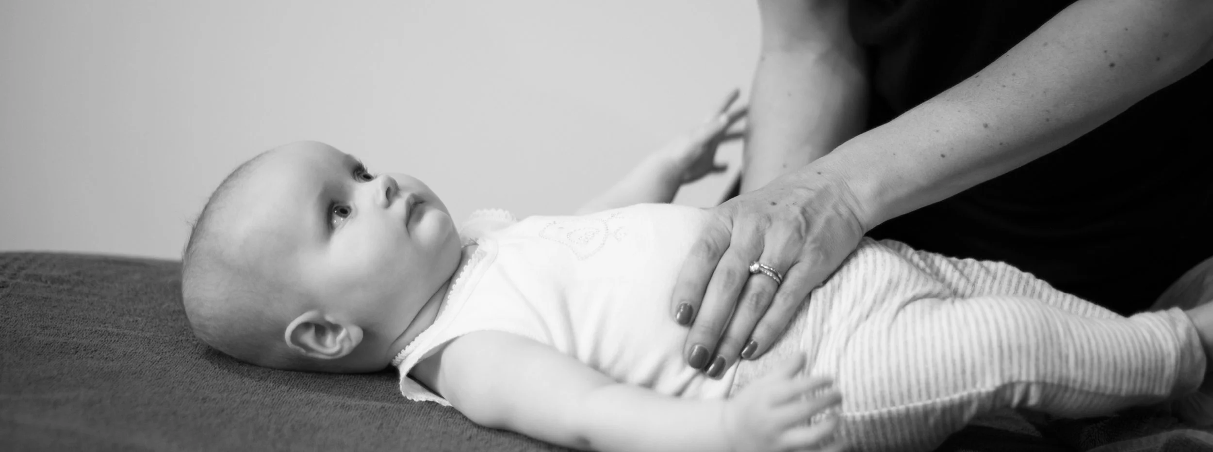 A baby lying on a table looking up at an adult's hand touching their chest, with the adult's arm extended and wearing a ring on their finger.