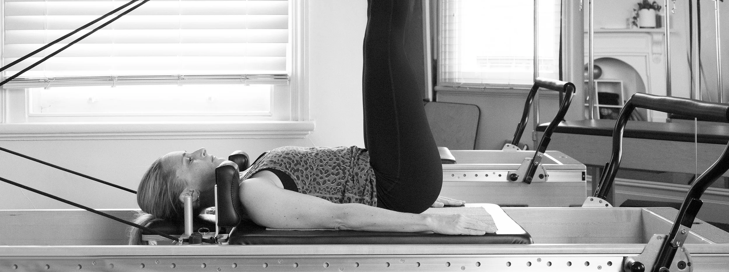 A woman is lying on her back on a Pilates reformer machine, with her legs raised straight up. She has a straight posture, with her head resting on the headrest, and her arms are relaxed at her sides. The room has large windows with blinds, and exercise equipment is visible in the background.