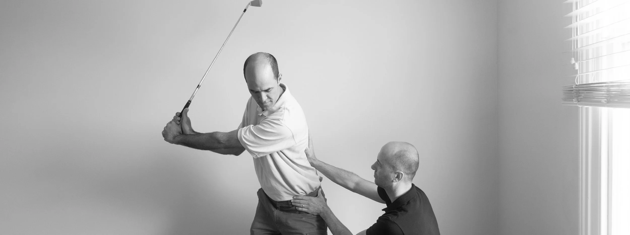 Two men, one standing and one kneeling, practicing golf swing together indoors. The man standing is holding a golf club and appears to be pretending to swing while the kneeling man, who is touching his back, offers guidance.