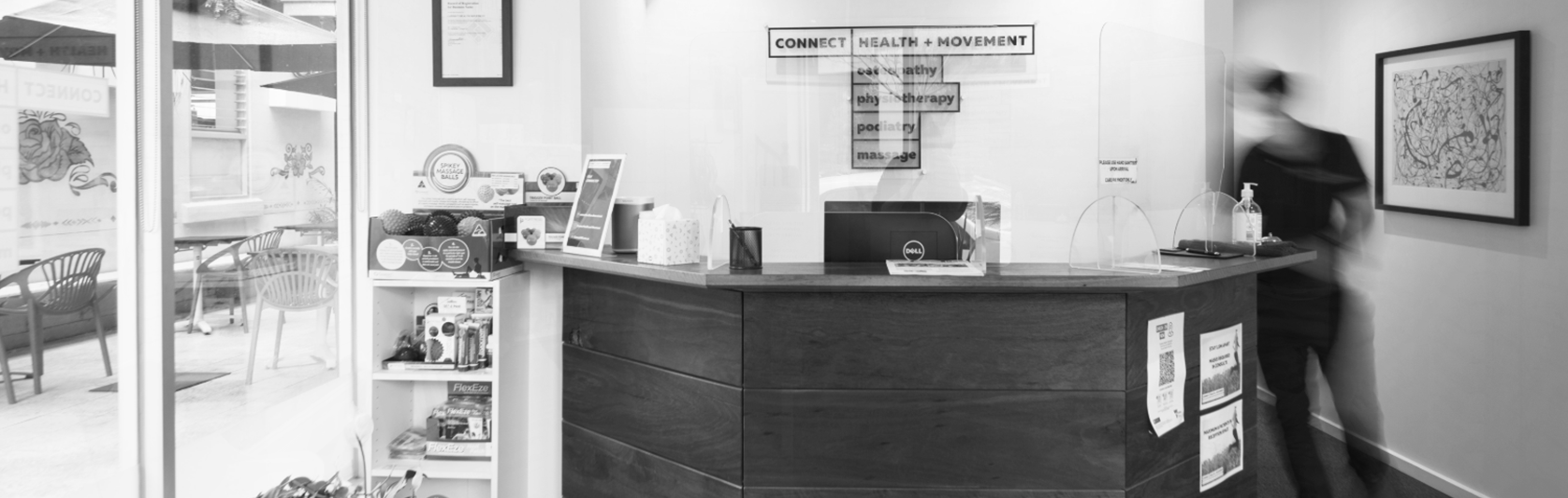 Photo of a reception desk in a health and movement clinic, with a person walking past and a blurred background showing artwork on the wall and a sitting area outside.